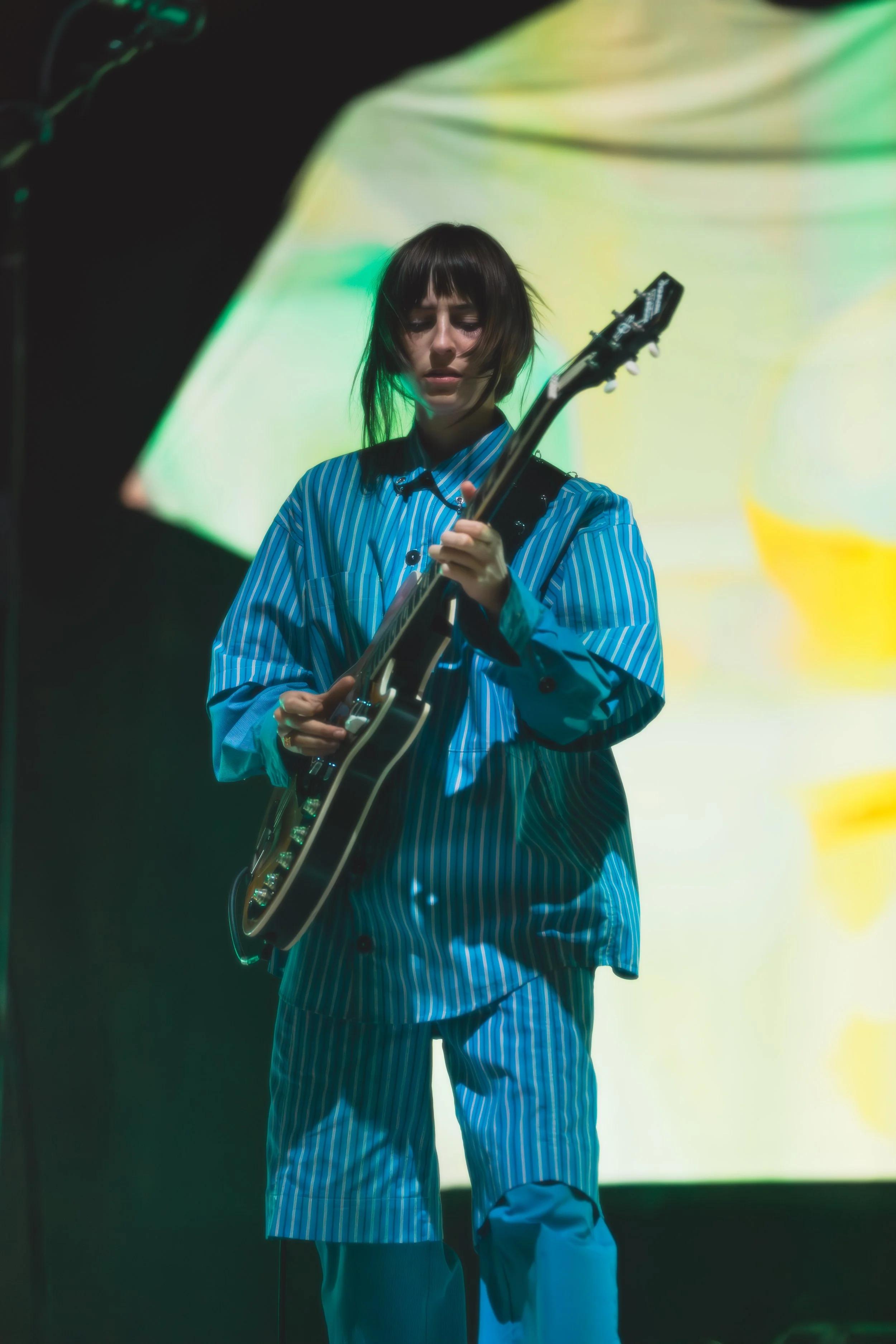 A woman with short dark hair wearing a blue striped shirt and matching shorts playing an electric guitar on stage with a colorful, abstract background projection.