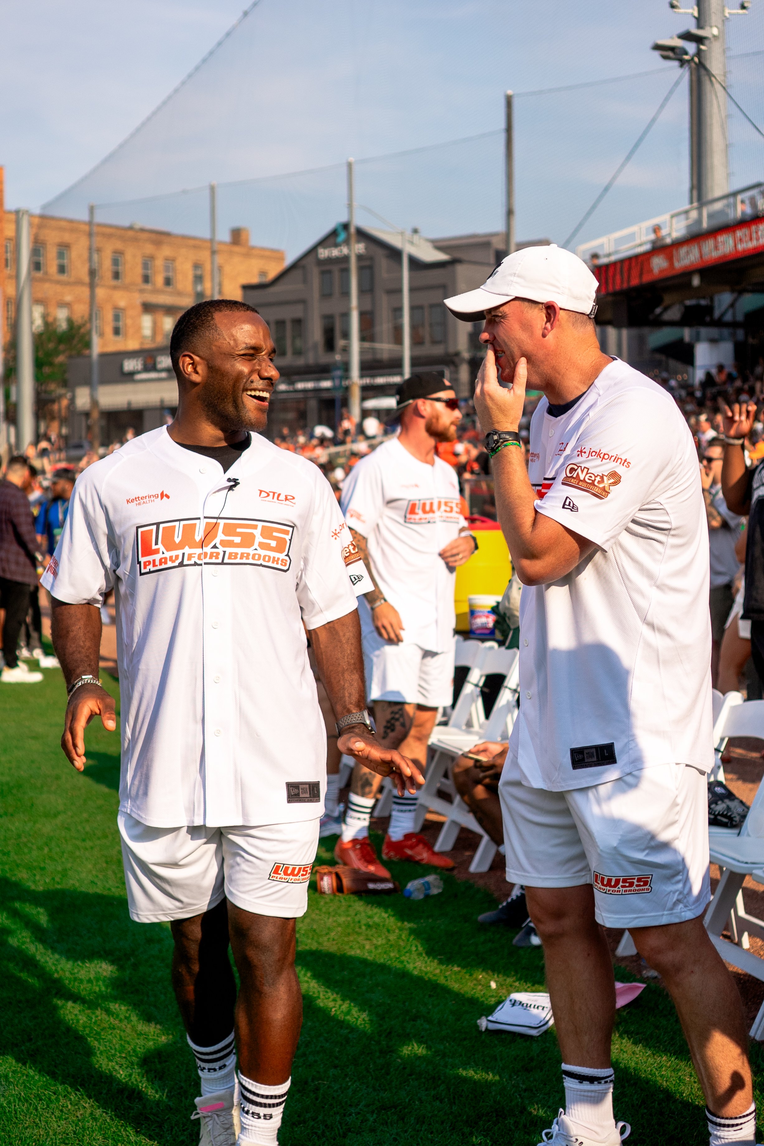 Two men dressed in white baseball jerseys and shorts talking and laughing during an outdoor event. One man is smiling widely, and the other has his hand on his face. There are more people and chairs in the background, and the setting appears to be a 