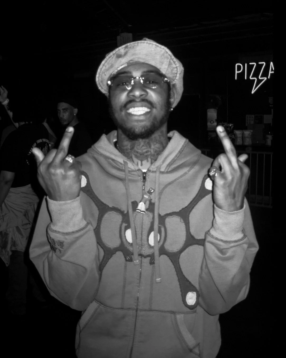 A man wearing glasses, a bucket hat, and a hoodie, making an obscene gesture with both hands, smiling. In the background, a neon sign says 'PIZZA' and there are other people.