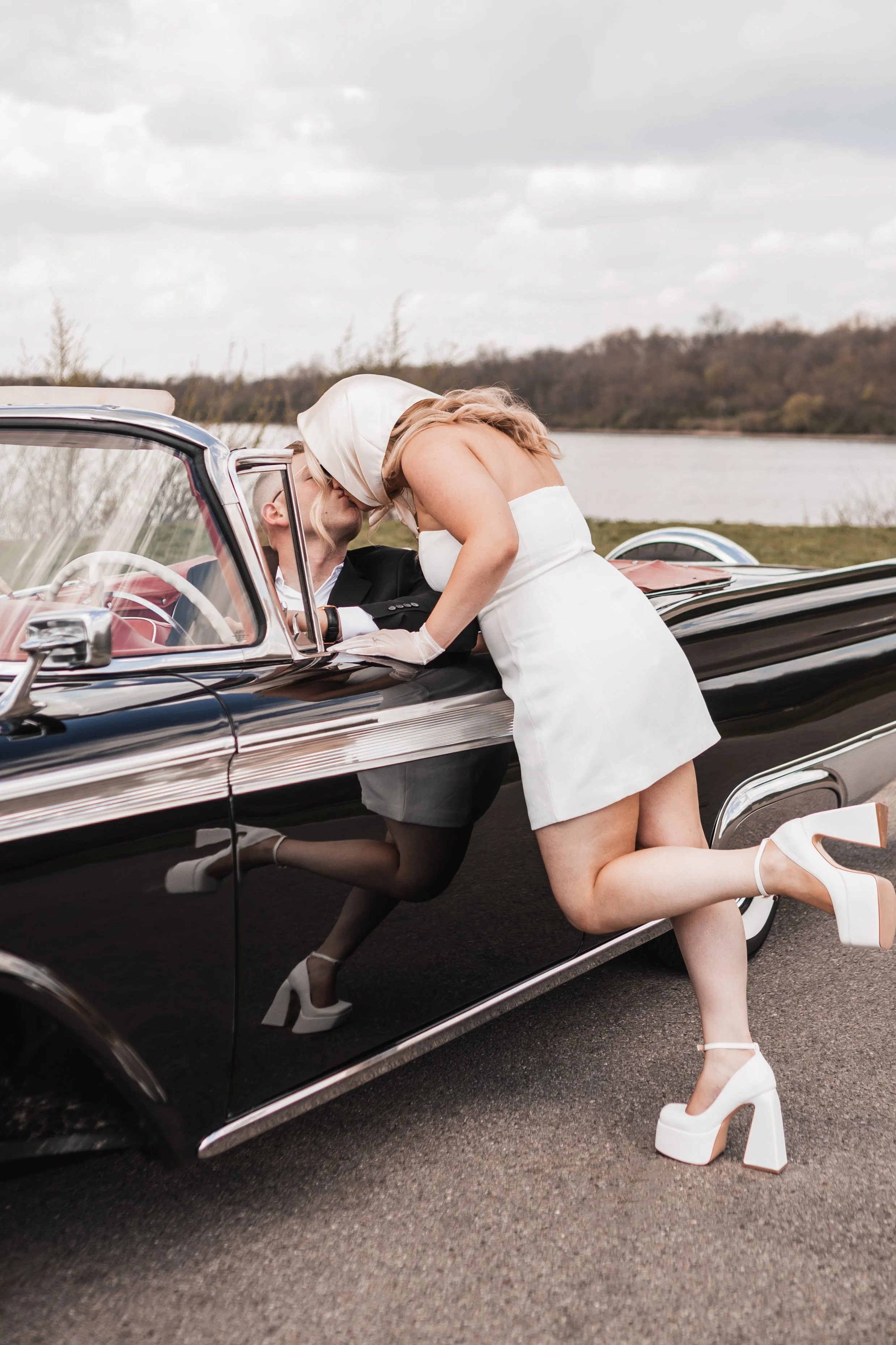 A woman in a white dress and white platform shoes kissing a man in a black suit while leaning into a classic black convertible car outdoors.
