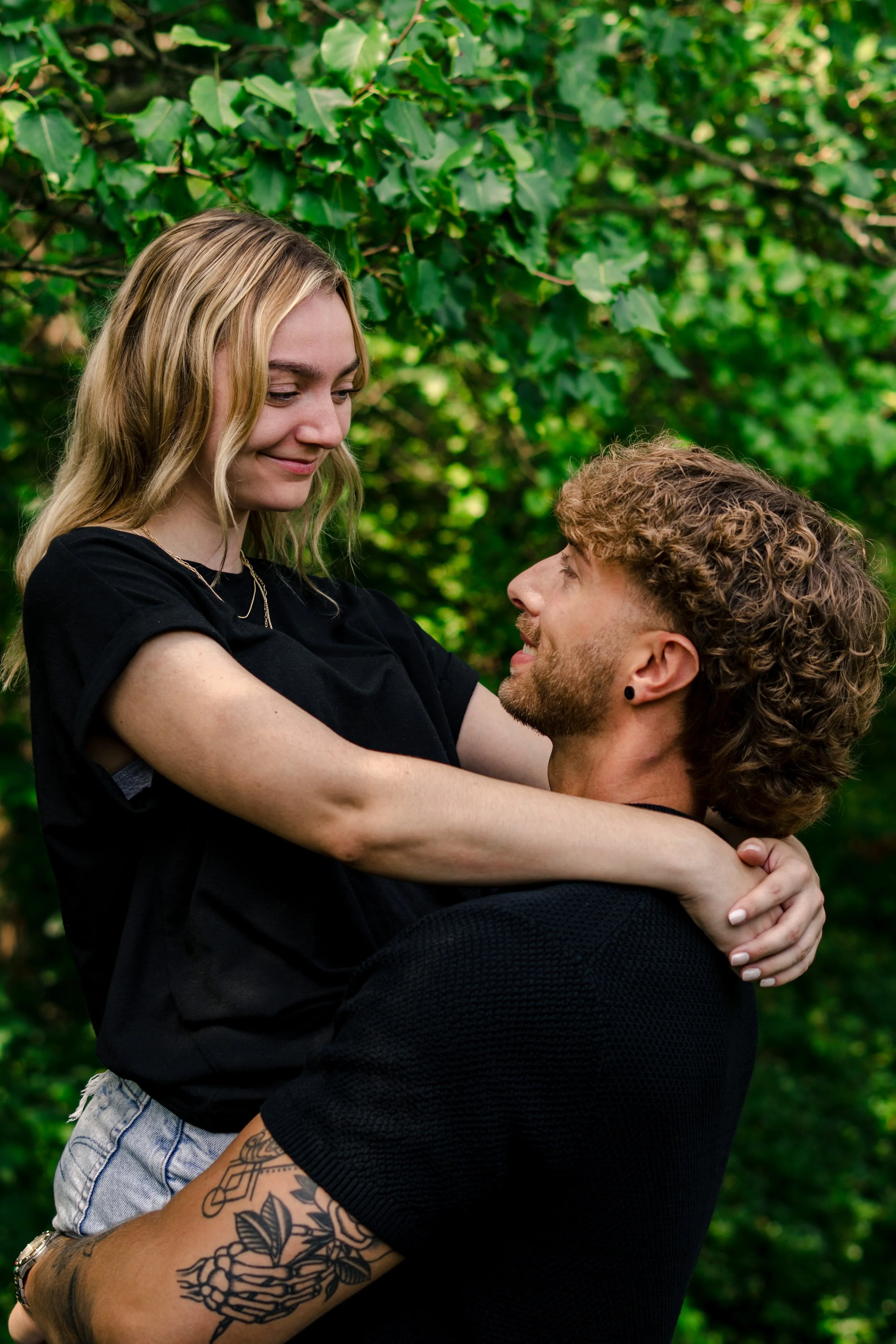 A young woman with blonde hair and a young man with curly hair and tattoos, engaging in a joyful embrace outdoors with green trees in the background.
