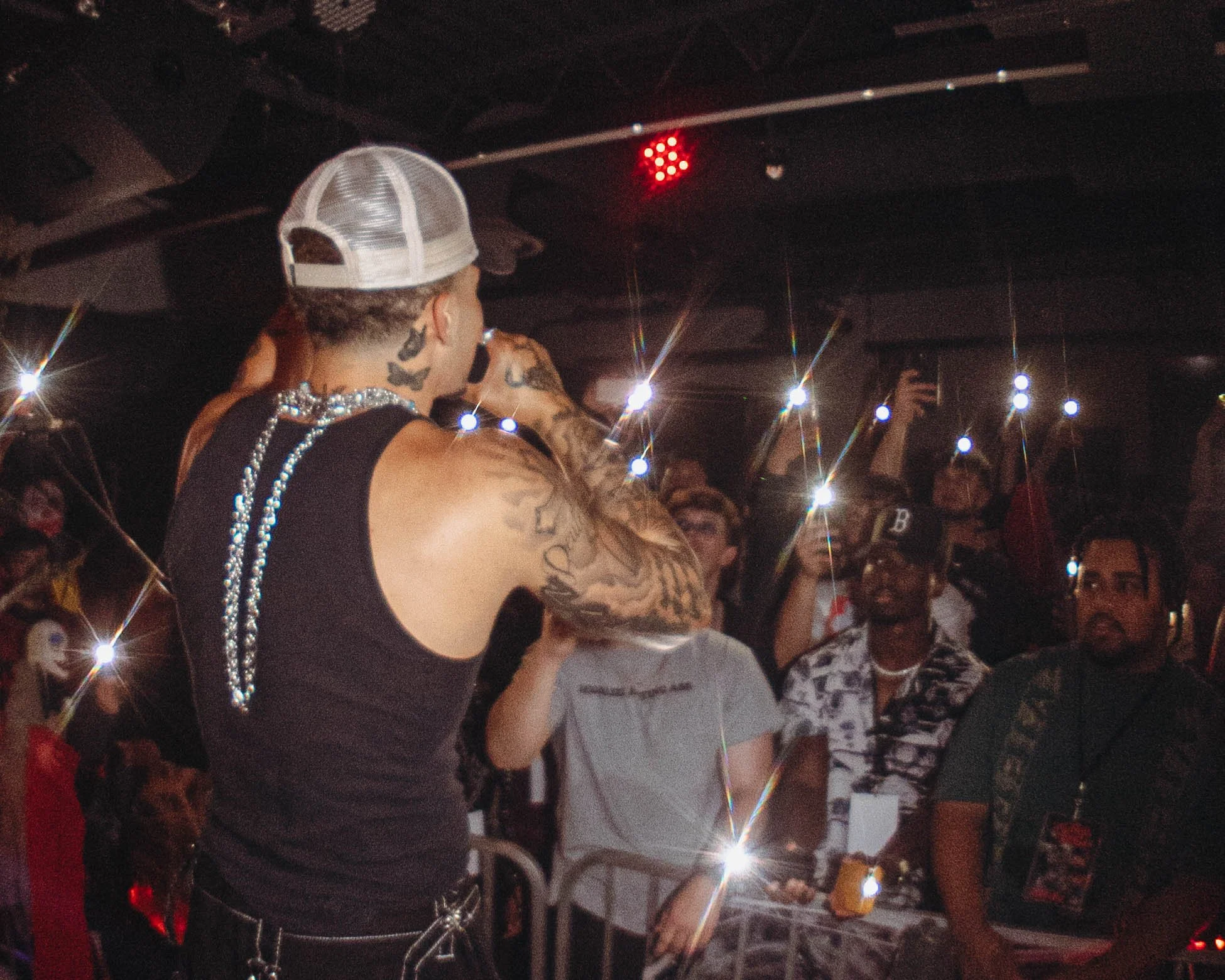 A performer with tattoos on arms, wearing a black sleeveless shirt, a cap, and a beaded necklace, holding a microphone on stage in front of an audience in a nightclub or concert venue.