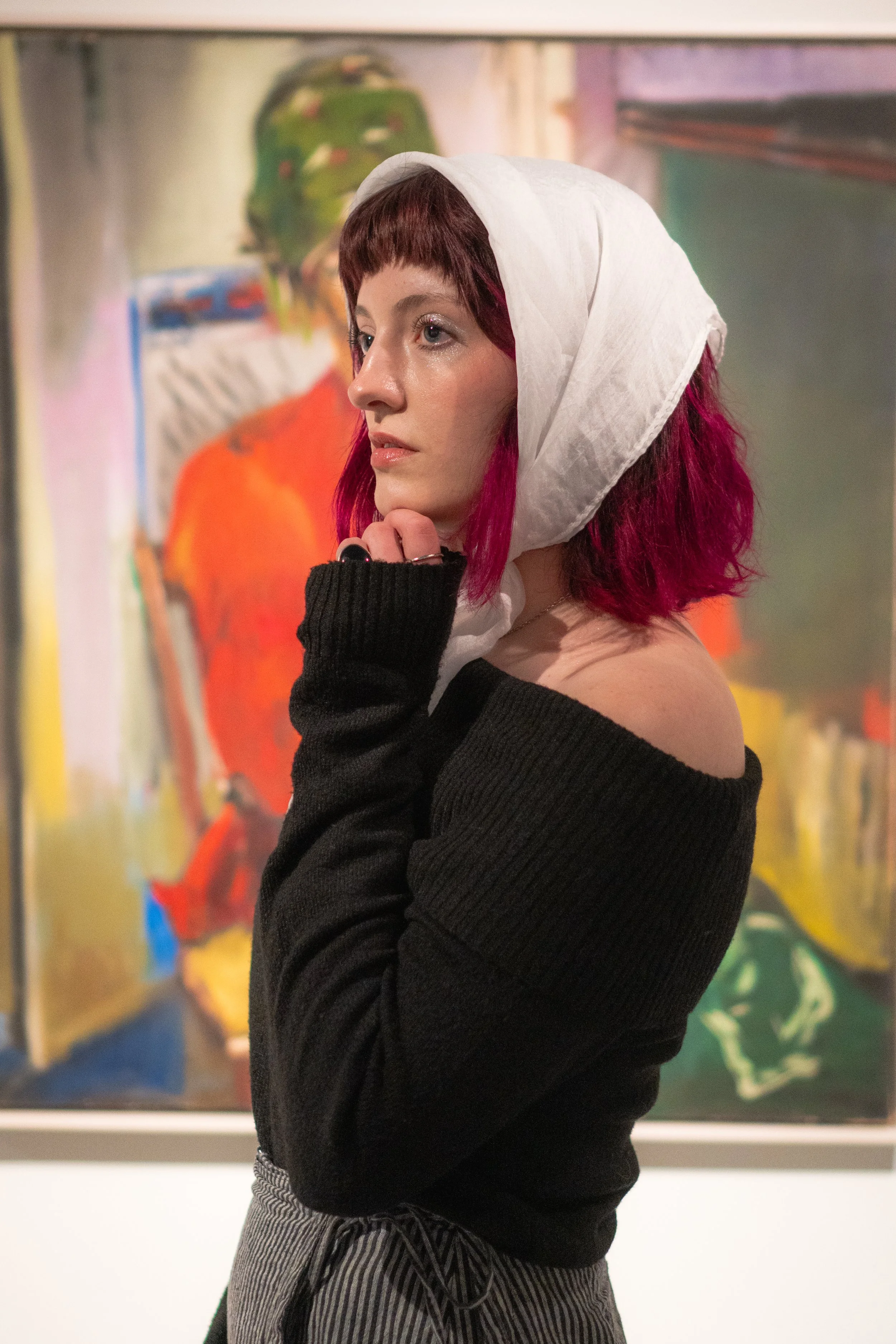 A young woman with pink hair and fair skin looking contemplative in an art gallery, wearing a white headscarf and black off-shoulder sweater, with a colorful abstract painting in the background.