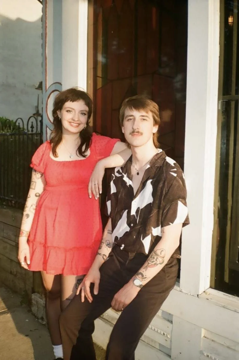 A young woman in a red dress with tattoos on her arms, standing next to a young man with tattoos on his arms, wearing a patterned short-sleeve shirt, outside near a building.