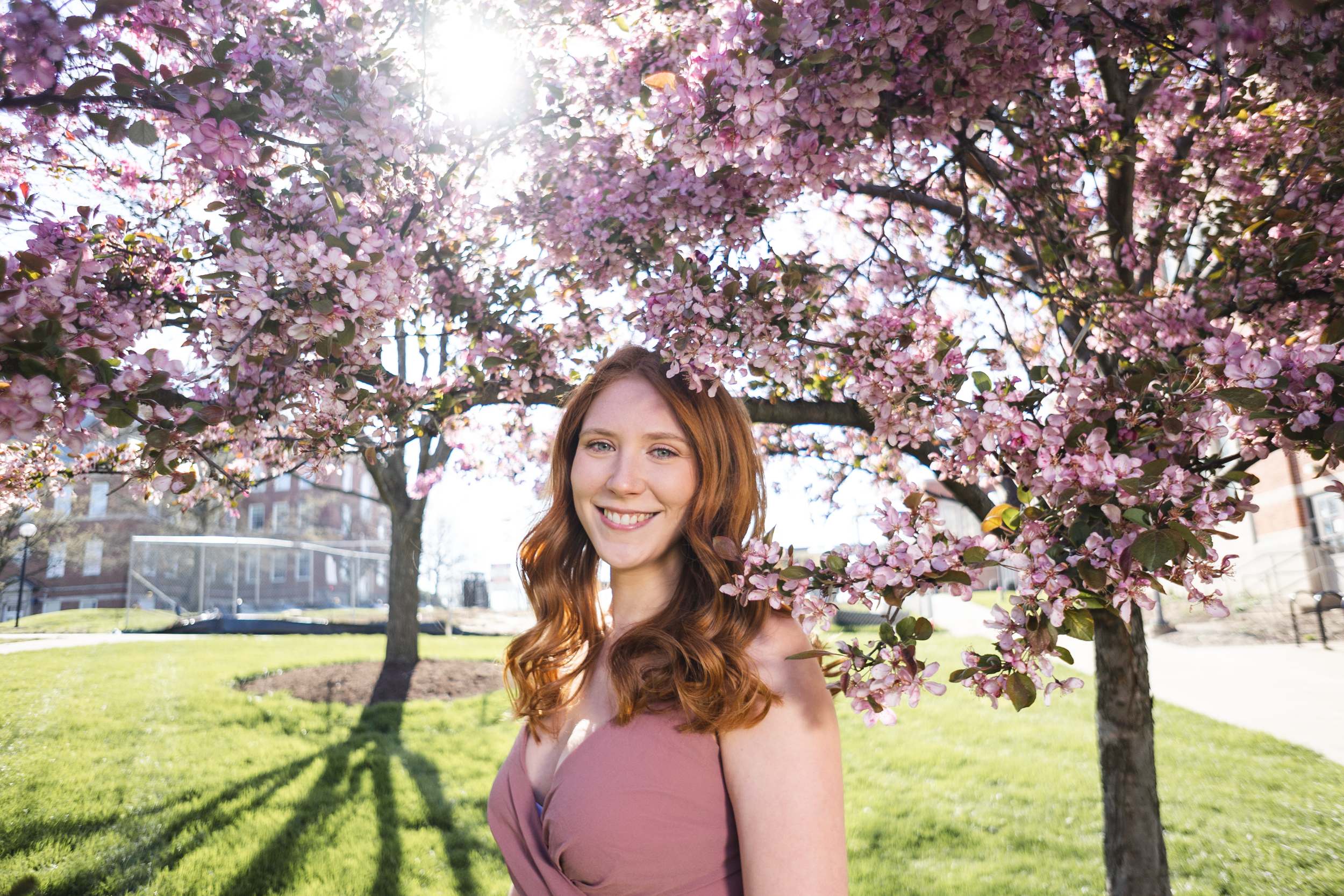 A young woman with red hair smiling in front of blooming pink cherry blossom trees on a sunny spring day in a park.