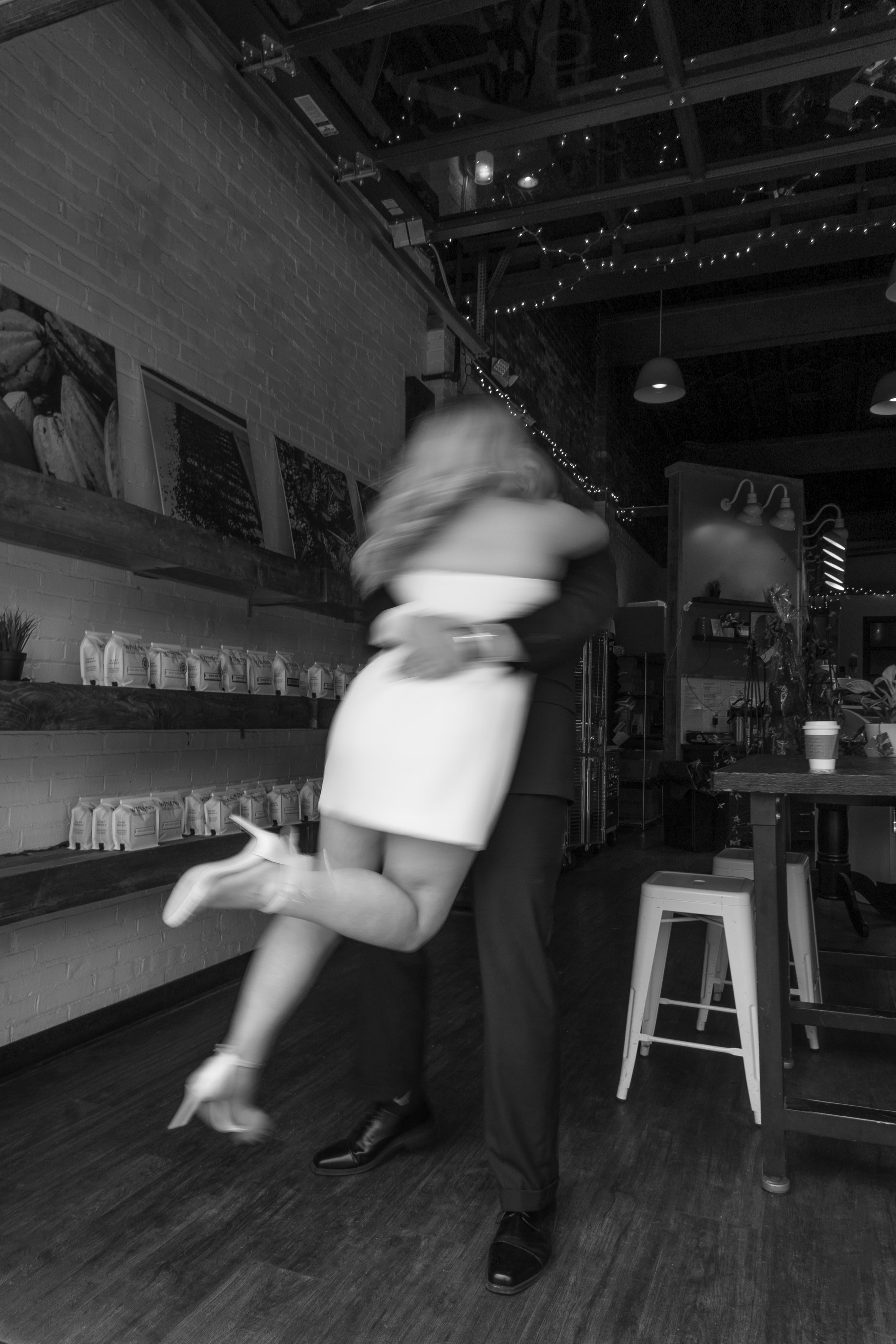 A couple dancing closely inside a cafe or restaurant, with one person lifting the other. The setting has exposed brick walls, pictures on display, and string lights hanging from the ceiling.