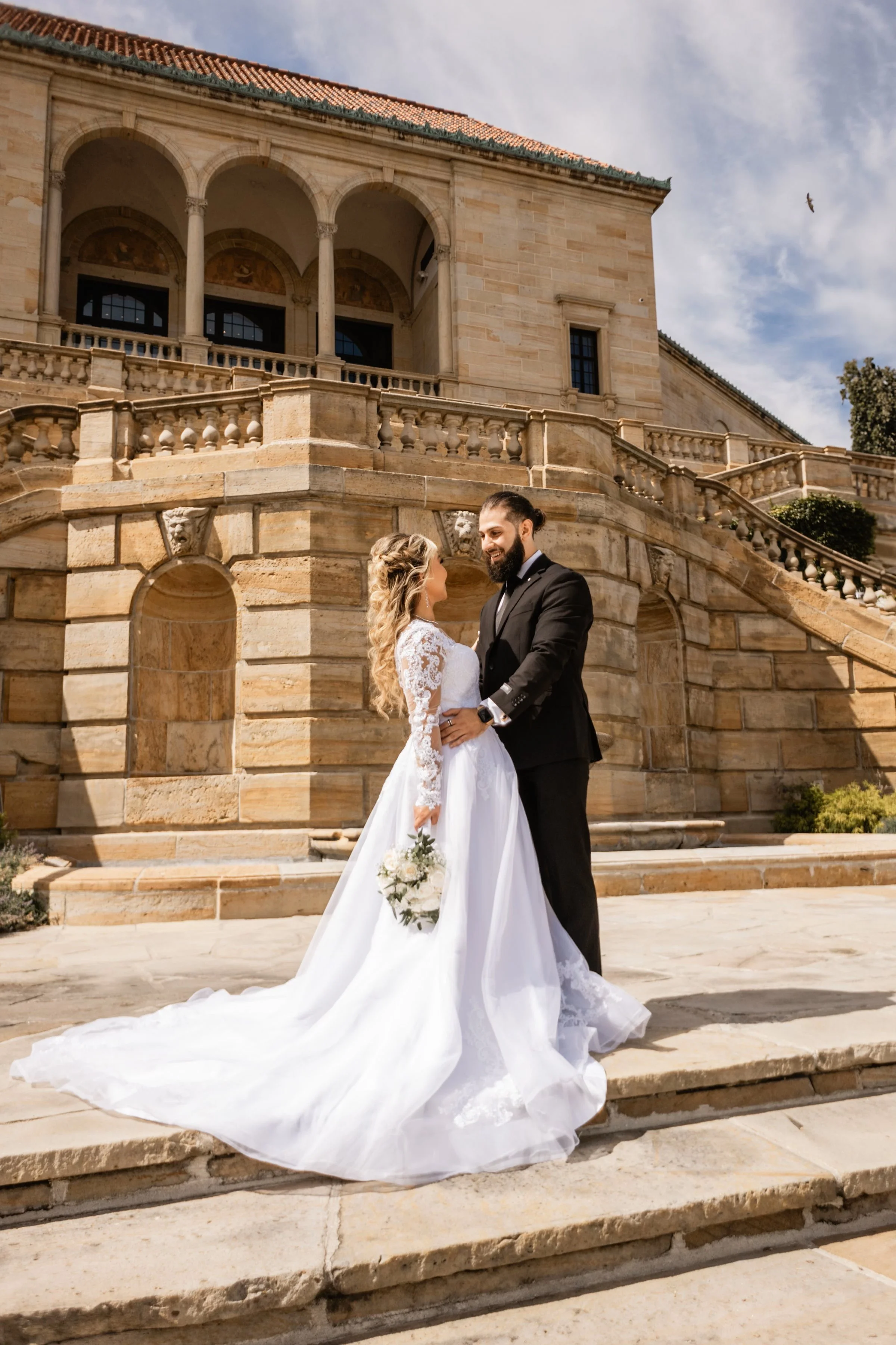 A bride and groom standing together outside in front of a historic stone building, holding hands and looking at each other, with the bride holding a bouquet of white flowers.