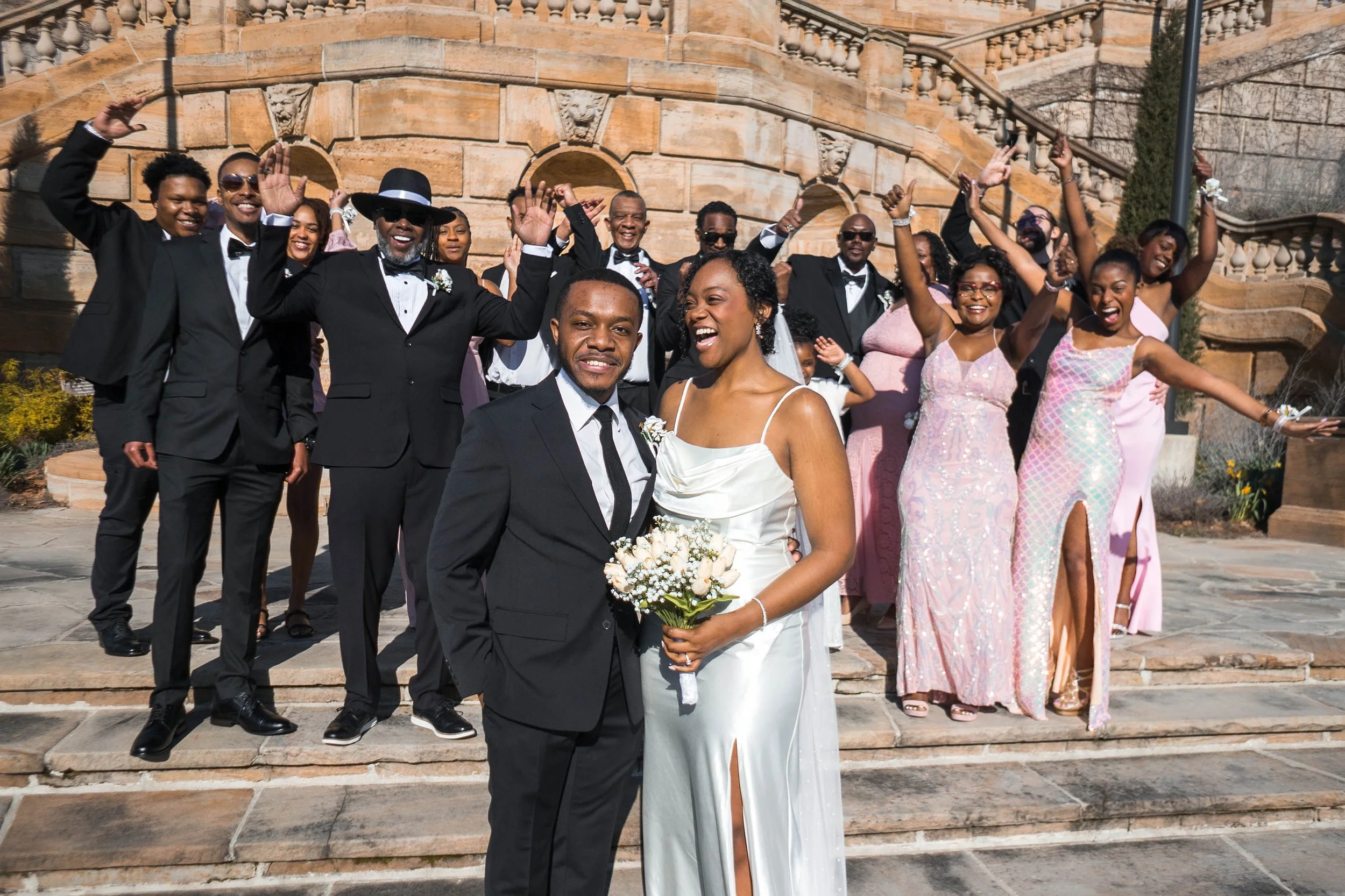 A wedding celebration on outdoor steps with a newlywed couple at the front, surrounded by friends and family dressed in formal attire, some with arms raised in celebration.