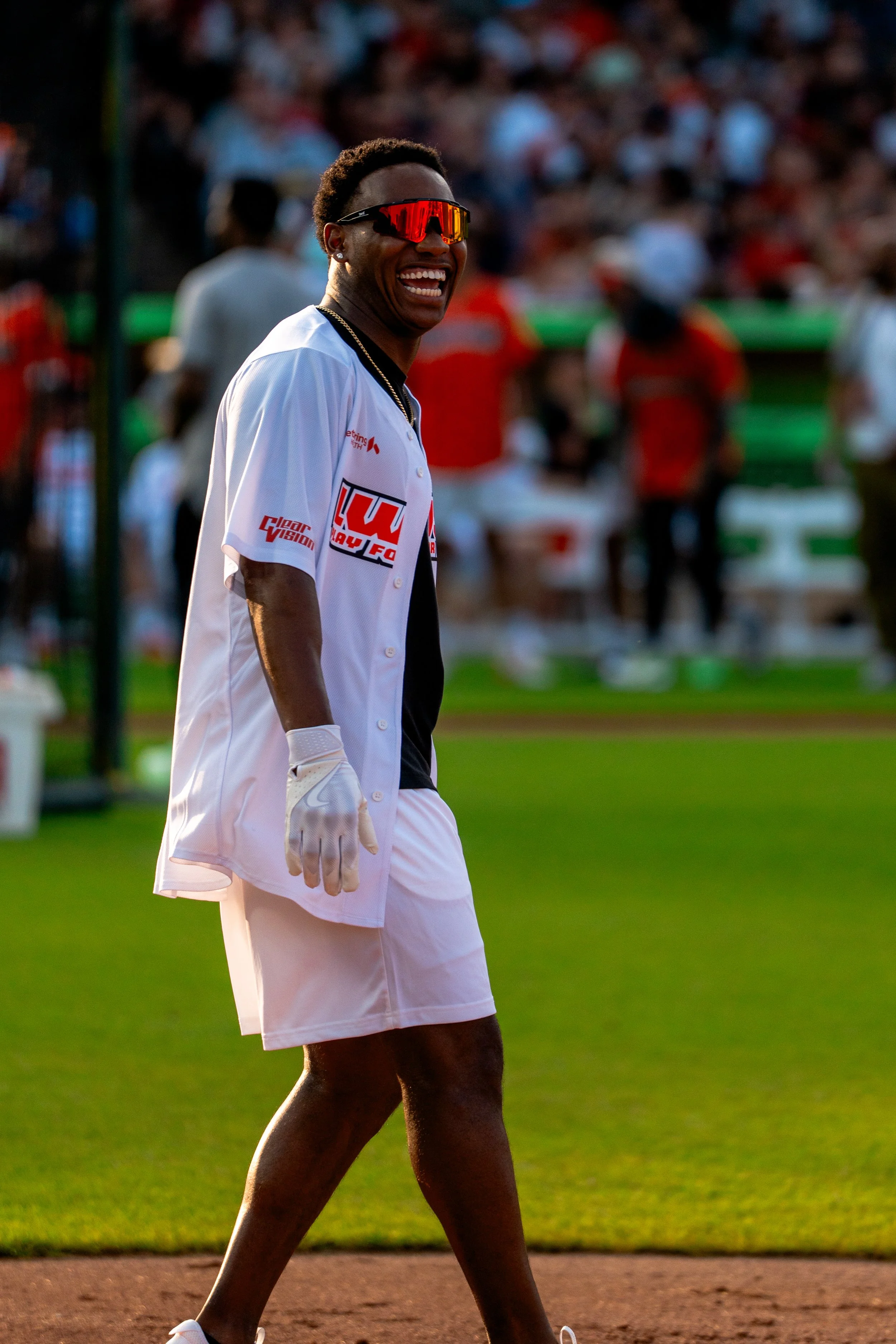 A smiling baseball player wearing a white jersey, black shirt underneath, white shorts, sunglasses, and a glove on his left hand on the field during a game.