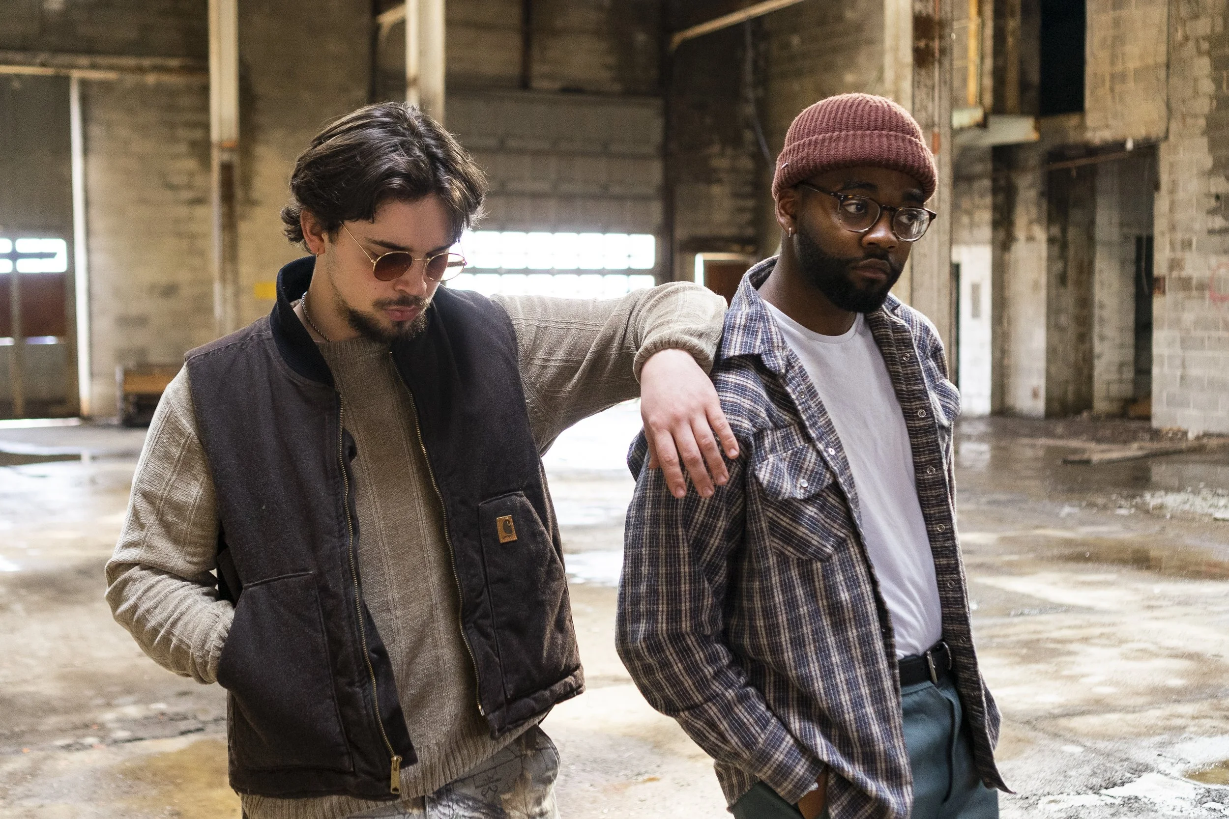 Two men walk inside an empty, industrial-style building with brick walls and large windows. One man has his arm over the shoulder of the other, and both appear somber or reflective.