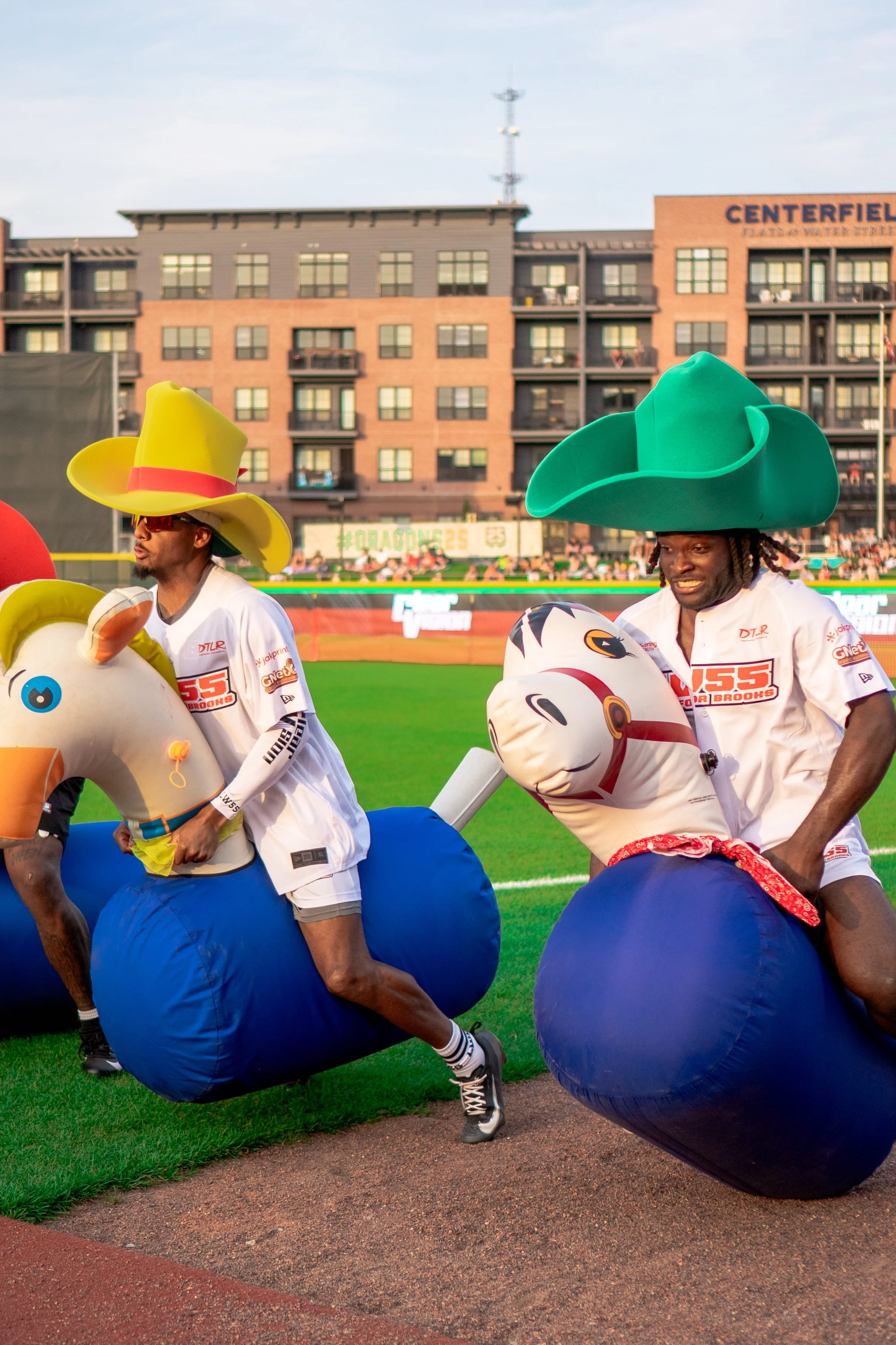 Two people in inflatable horse costumes and oversized cowboy hats are on a baseball field during a game, with other spectators in the background.