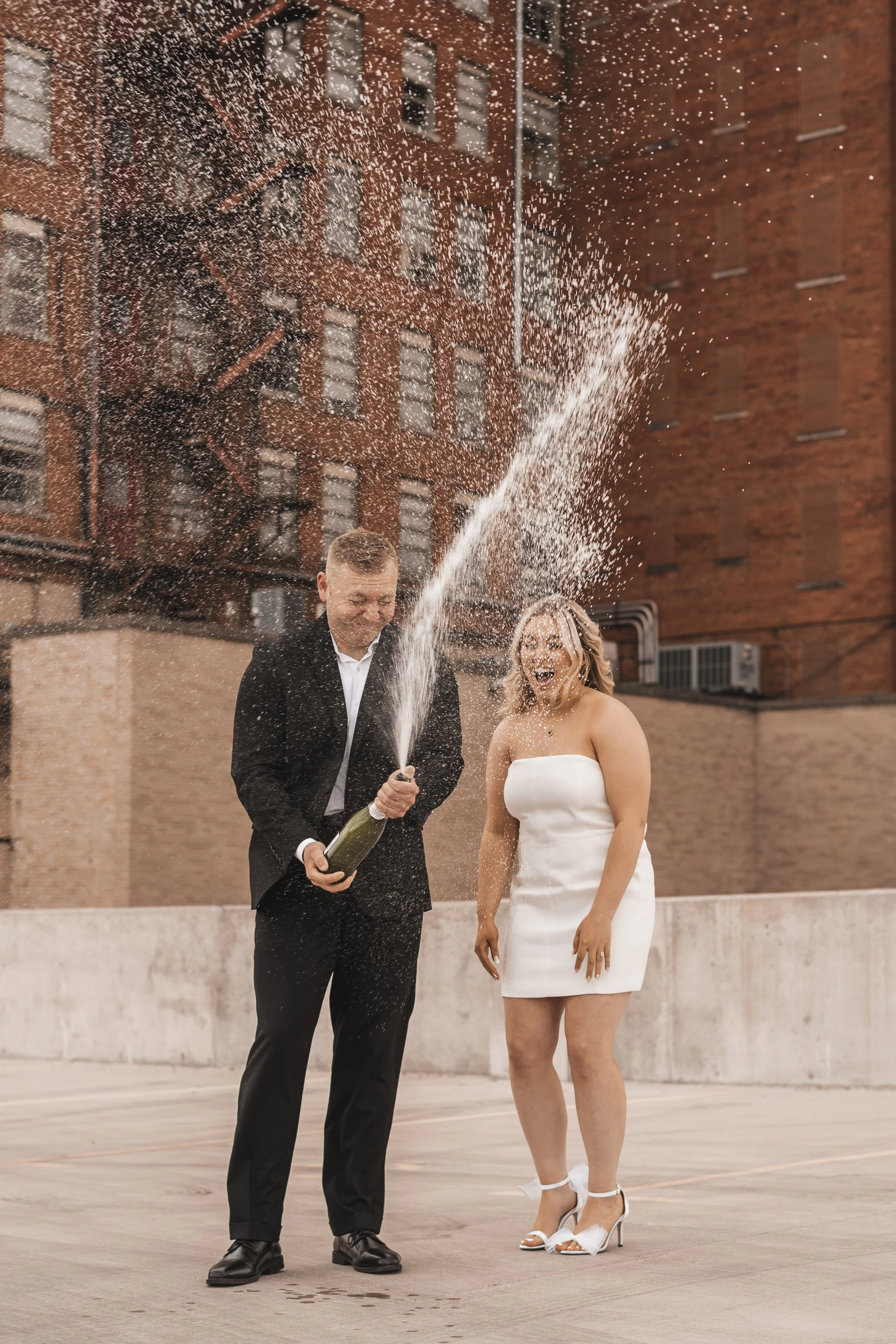 A man in a black suit and a woman in a white dress celebrating on a rooftop with a champagne bottle, opening it as champagne sprays out, both smiling and laughing. Background shows tall brick buildings.