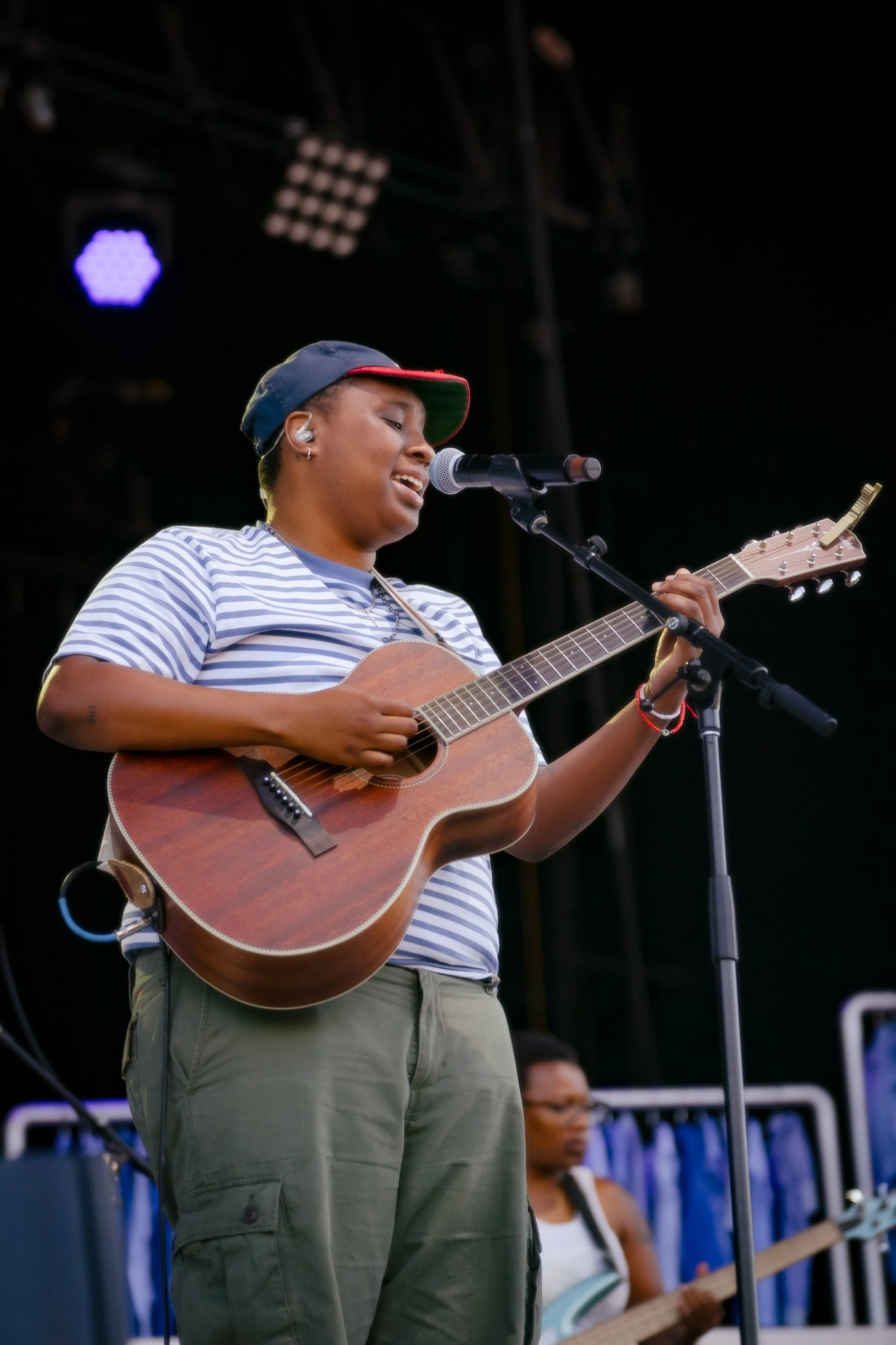 A young woman with short hair, wearing a striped t-shirt, green cargo pants, a blue cap with red trim, and earbuds, is performing on stage with an acoustic guitar, singing into a microphone.