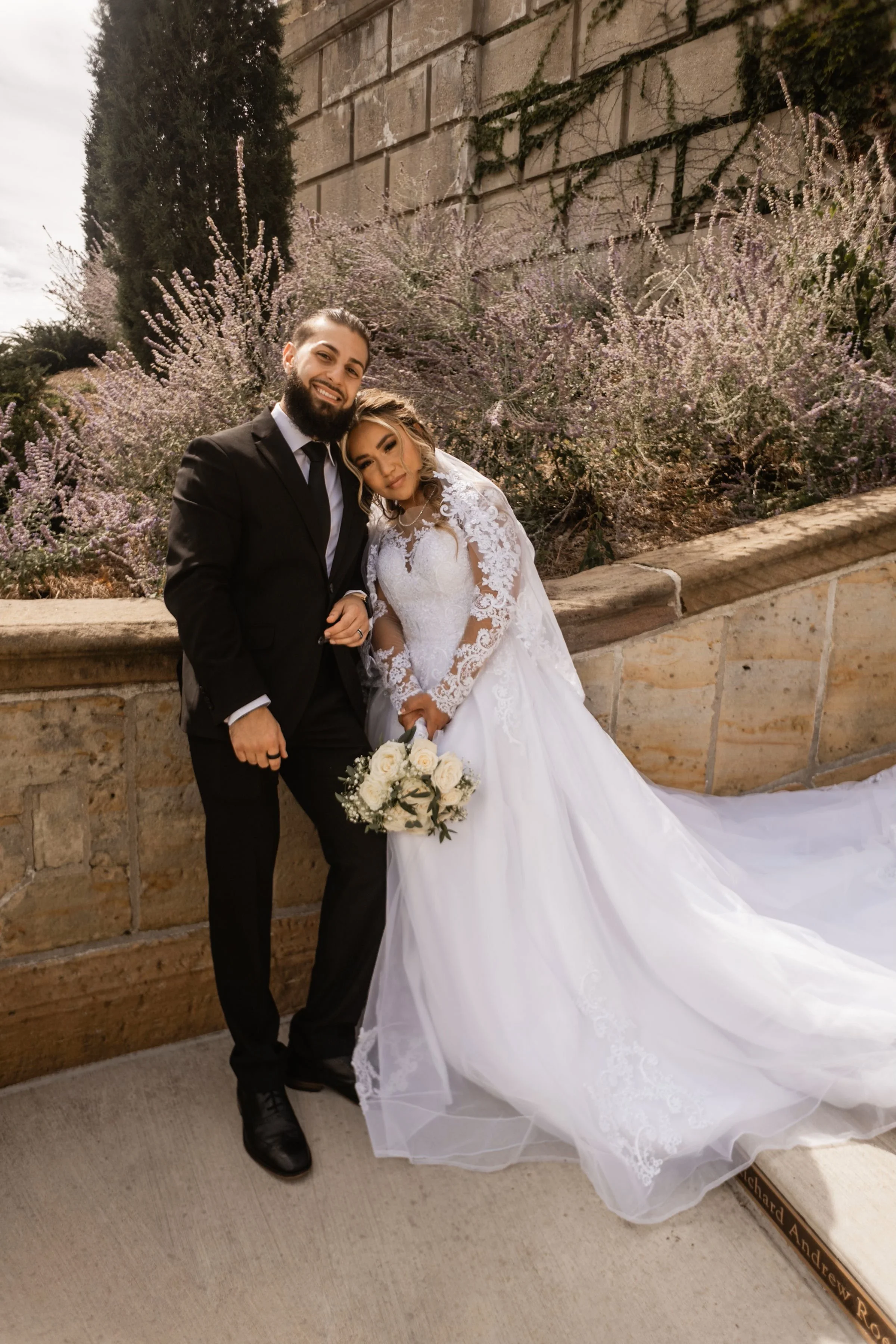 A bride and groom posing together outdoors, with the bride in a white lace wedding gown holding a bouquet and the groom in a black suit, against a backdrop of flowering bushes and stone wall.