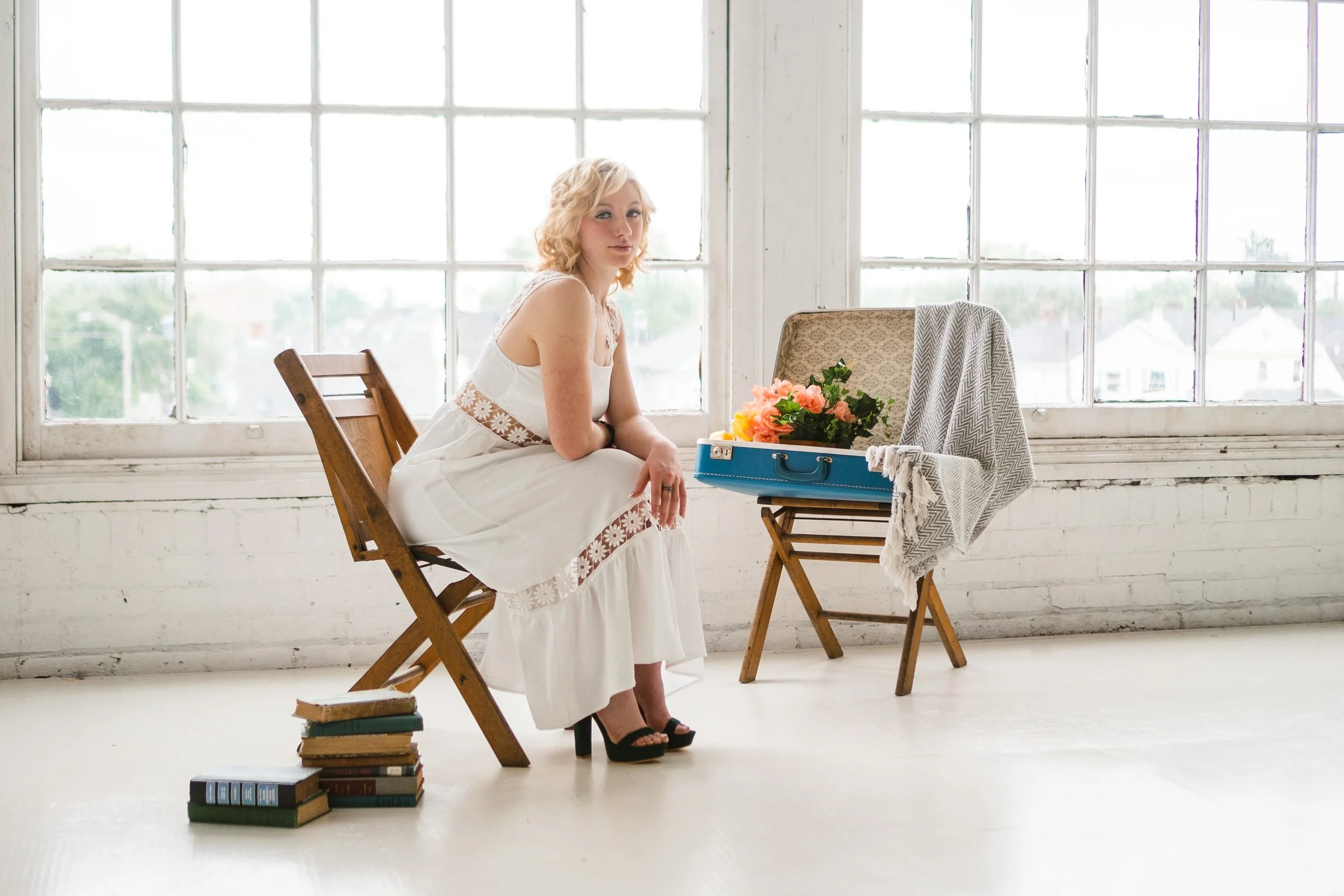 A woman in a white dress sitting on a wooden chair next to a small table with a blue suitcase containing pink flowers, with a window in the background.
