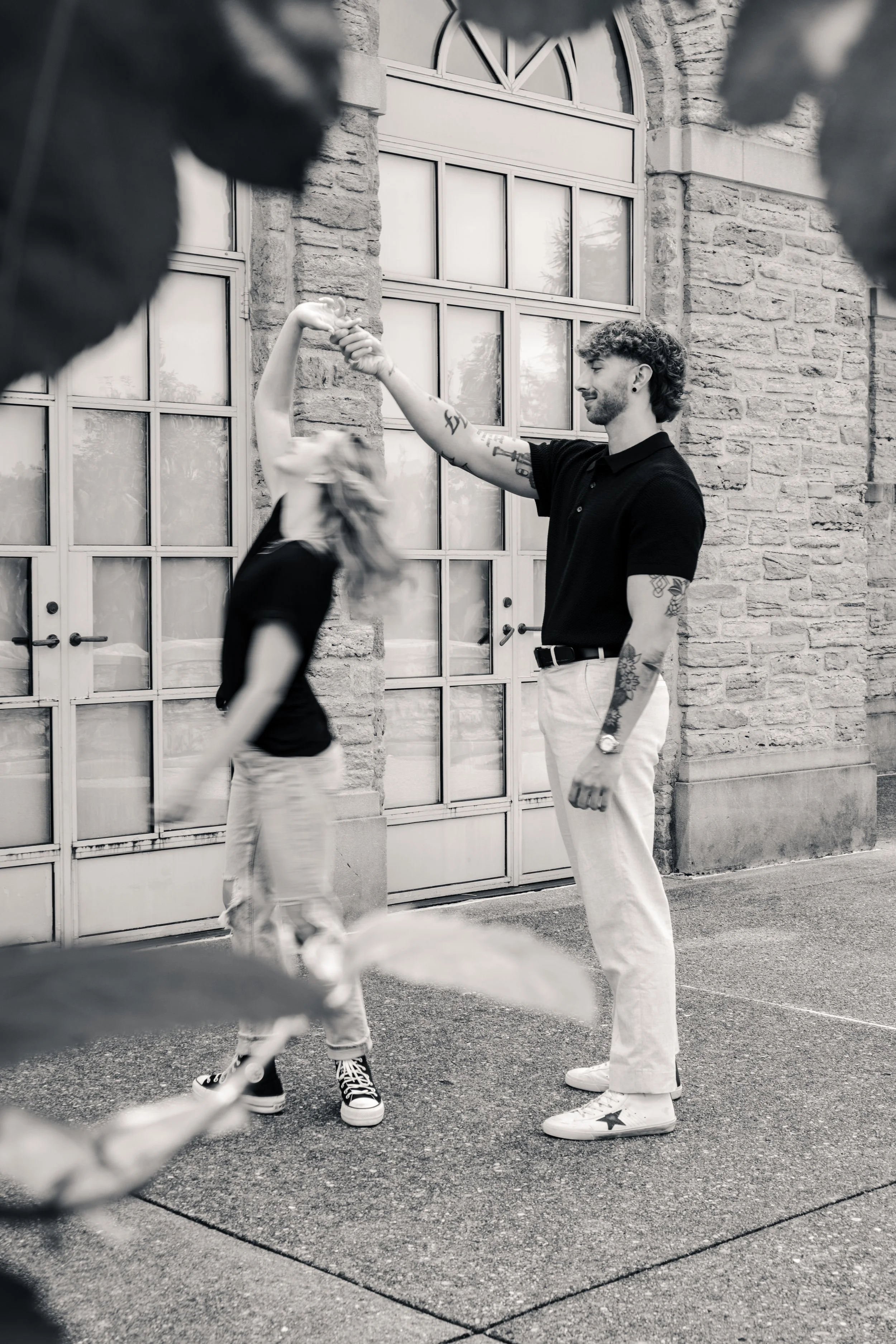 A man with tattoos and curly hair is dancing outdoors with a woman with long hair, both are wearing black shirts and light-colored pants, framed by some leaves in the foreground, in front of a brick building with large windows.