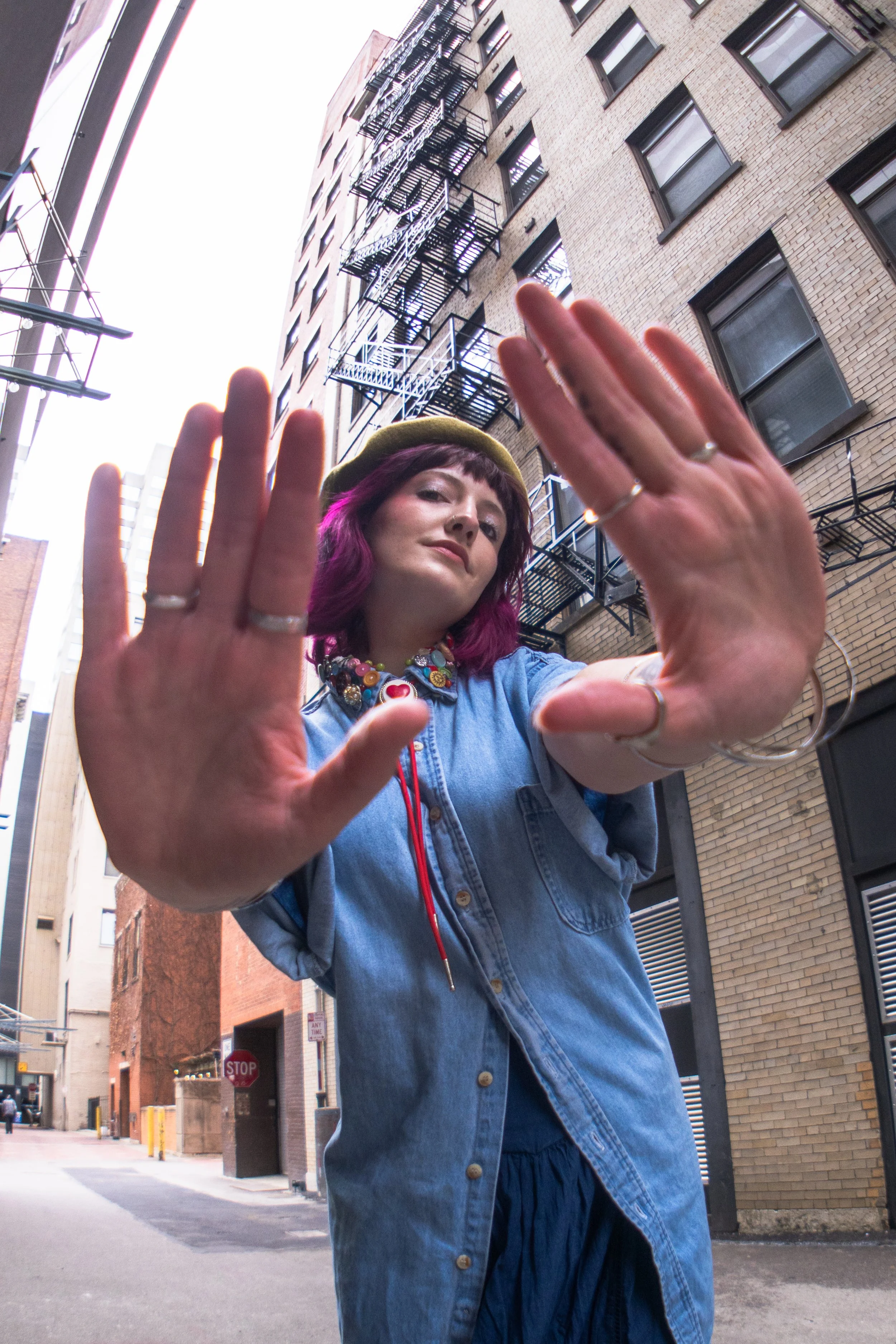 A woman with purple hair wearing a yellow beret, a denim shirt, and colorful jewelry, holds her hands up towards the camera in an alleyway surrounded by brick buildings.