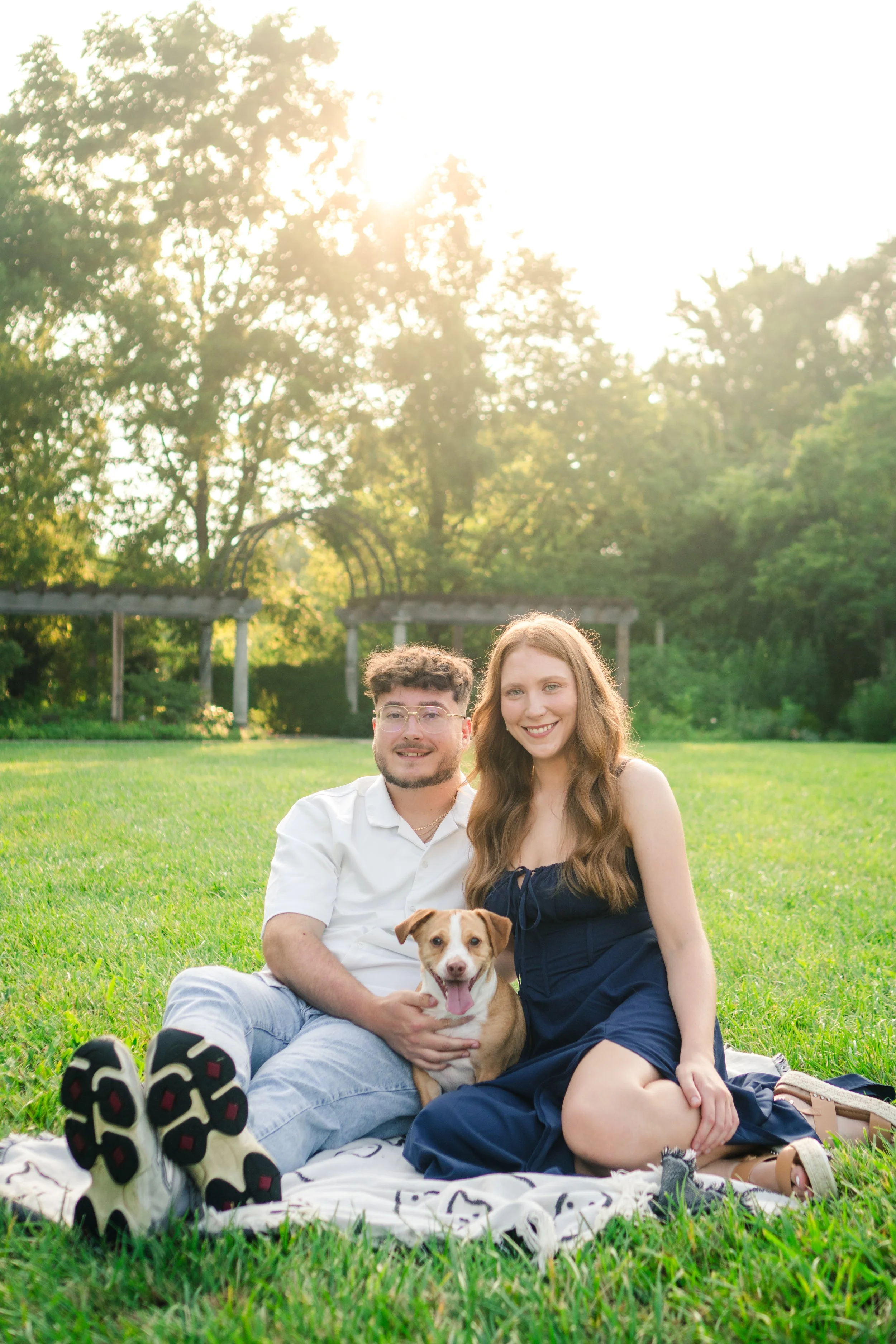 A young couple with a dog sitting on a blanket in a park during sunset, surrounded by green grass and trees.