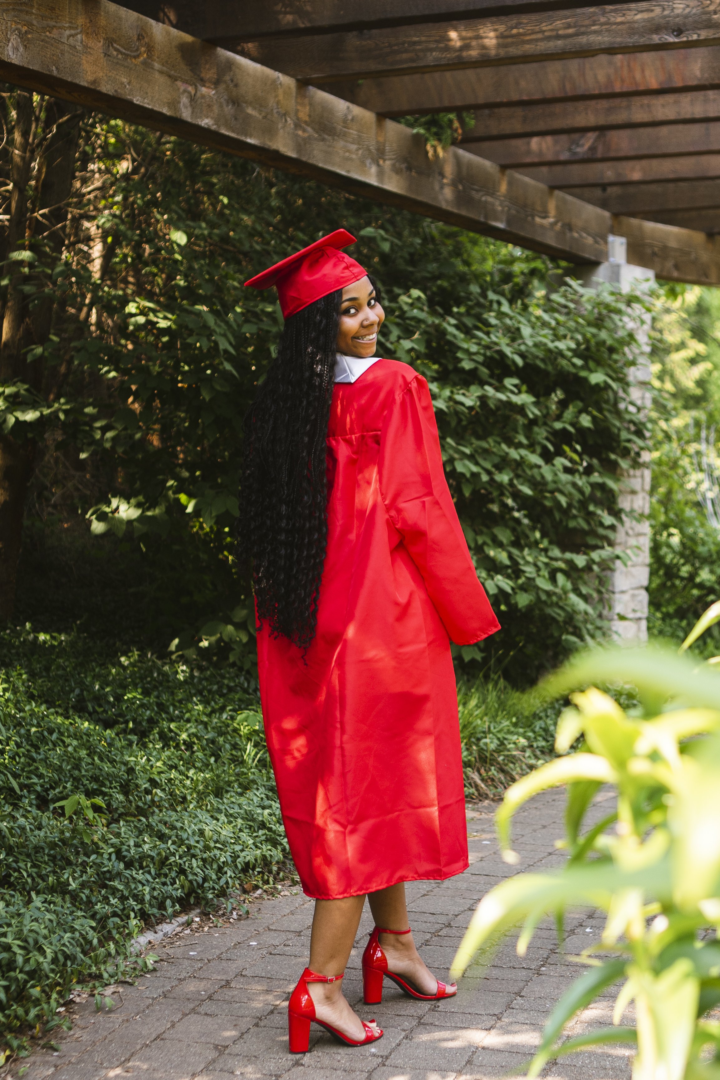 Young woman in a red graduation gown and cap, smiling and turning back, standing on a brick pathway surrounded by greenery.