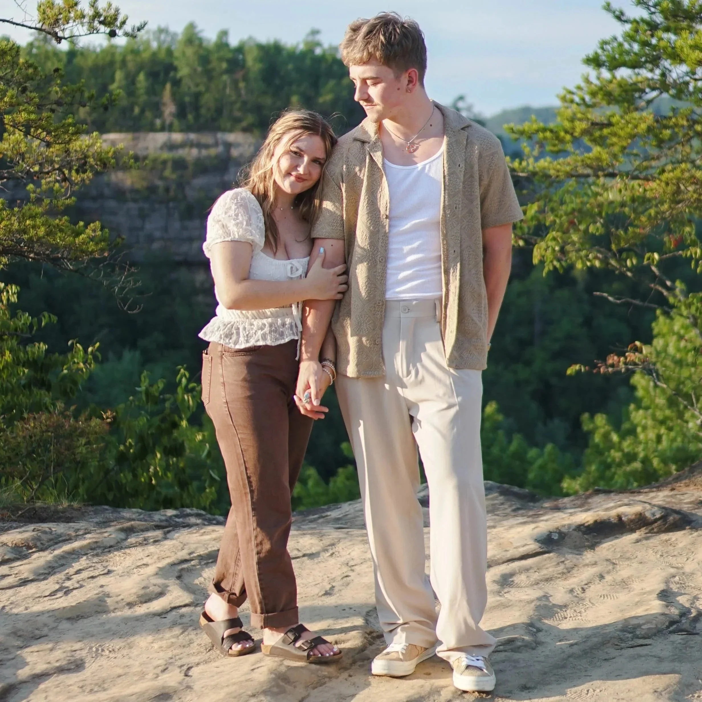 A young couple standing on a rocky ledge outdoors with green trees and hills in the background. The woman is holding the man's hand and leaning on his shoulder, both smiling softly.
