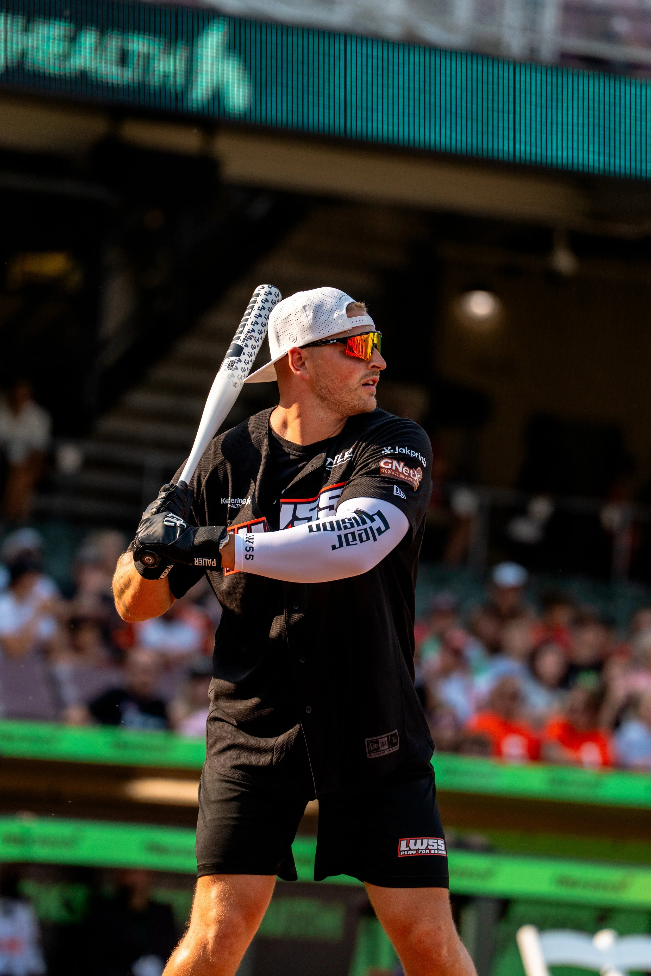 A baseball player in black attire, wearing a white cap backwards and reflective sunglasses, is preparing to swing a baseball bat during a game.