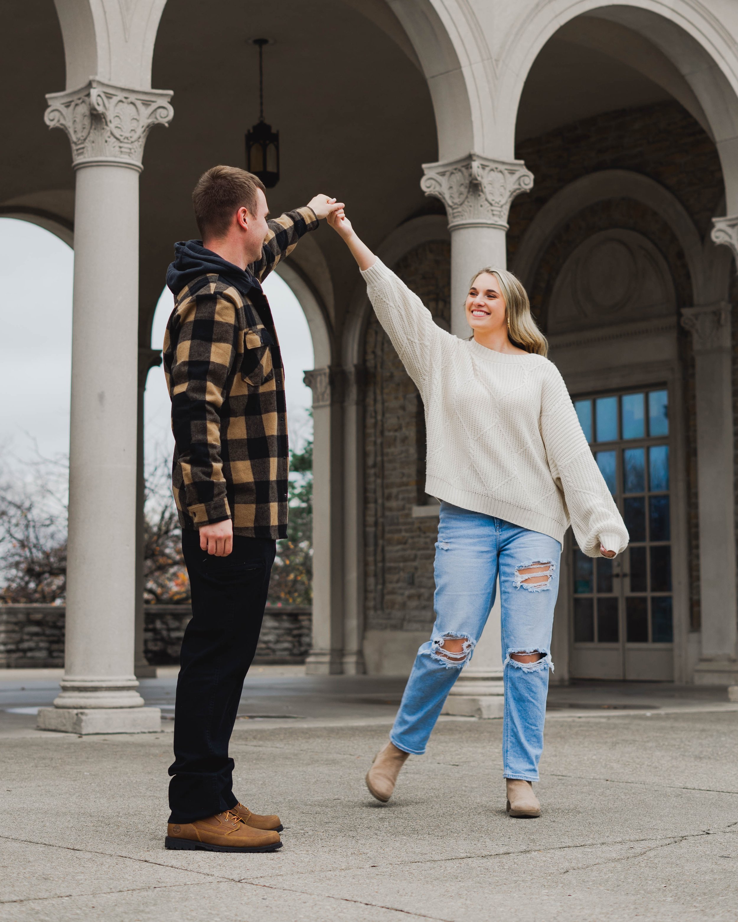 A young woman and man dancing outdoors on a stone patio with arches and columns in the background. The woman is wearing a white sweater, ripped jeans, and beige ankle boots, smiling as she twirls the man. The man is wearing a checkered jacket, dark p