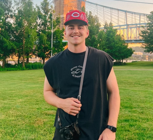 Young man smiling outdoors in a park with trees and a bridge in the background, wearing a red Cincinnati Reds cap, black sleeveless shirt, and a camera hanging from his neck.