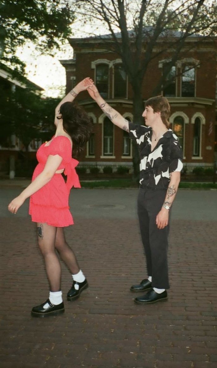 A young couple dancing on a brick street in front of an old brick building with large windows and trees. The woman is wearing a bright red dress, black fishnet stockings, and black shoes, while the man is dressed in a black and white shirt, black pan
