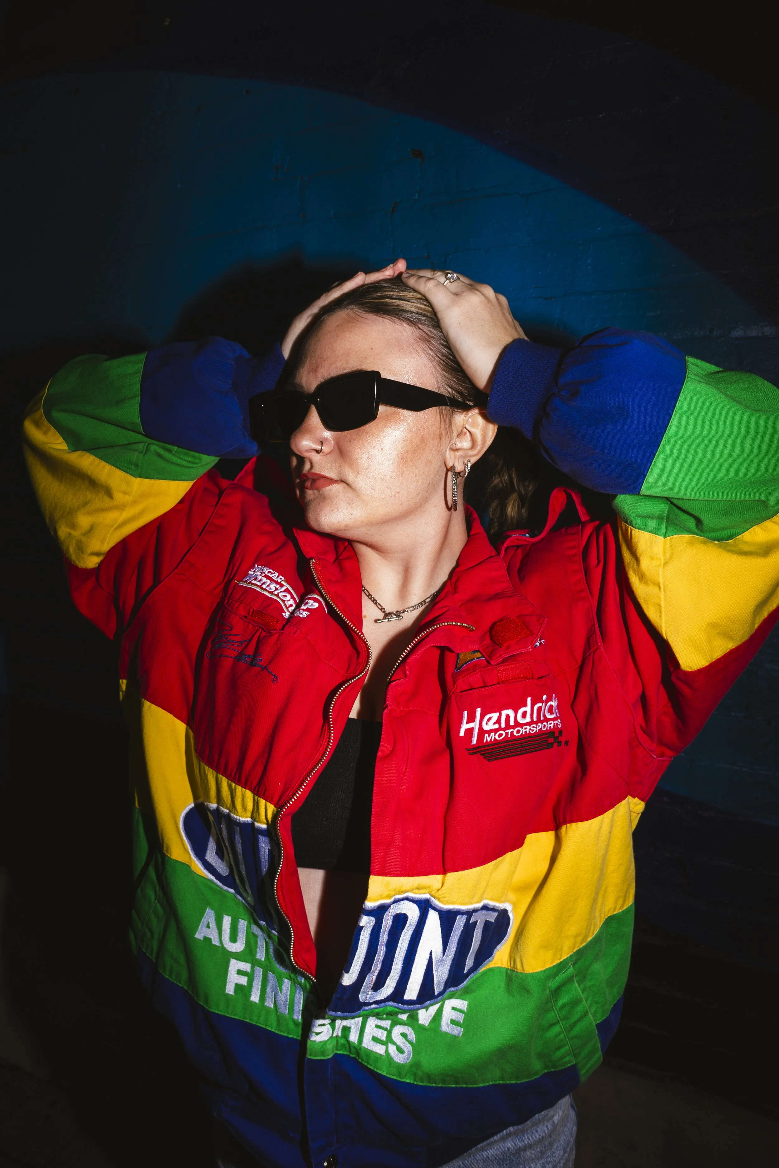 Person wearing a multicolored racing jacket with logos, black sunglasses, and earrings, posing with hands on head against a dark background.