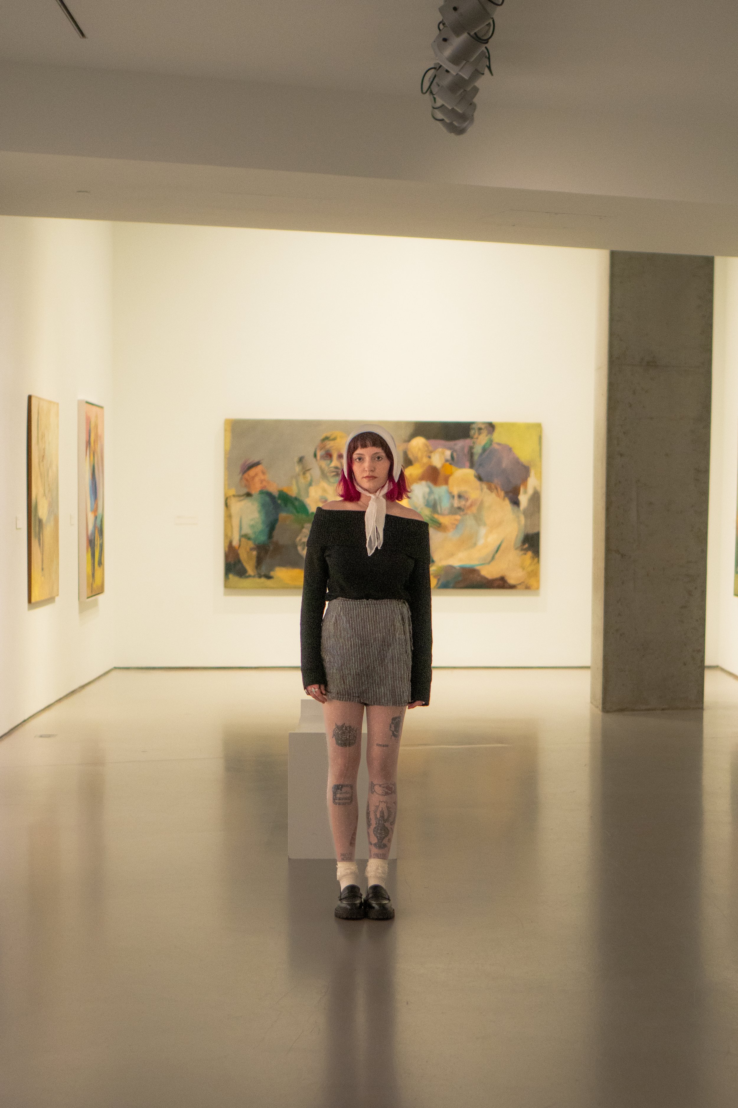 A young woman with pink hair and tattoos on her legs standing in an art gallery, with abstract paintings on the walls behind her.