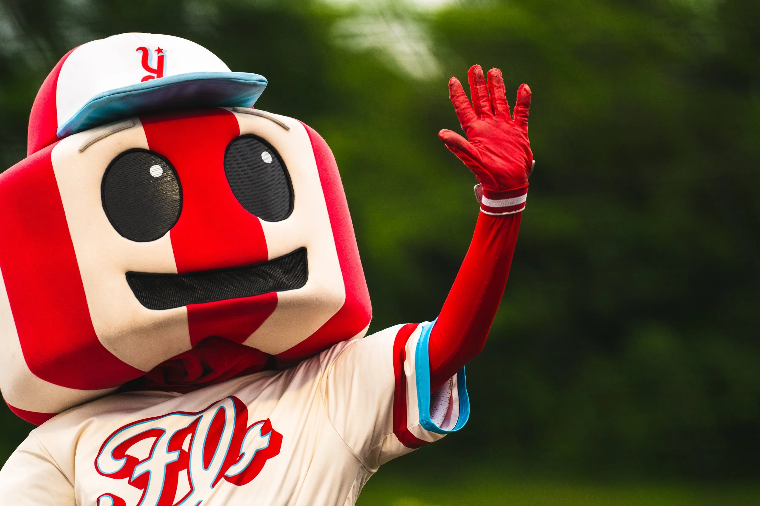 Mascot with a red and white striped face, large black eyes, and a big black smile, wearing a baseball uniform with 'FOS' on the front and a red, white, and blue cap, waving with a red-gloved hand.