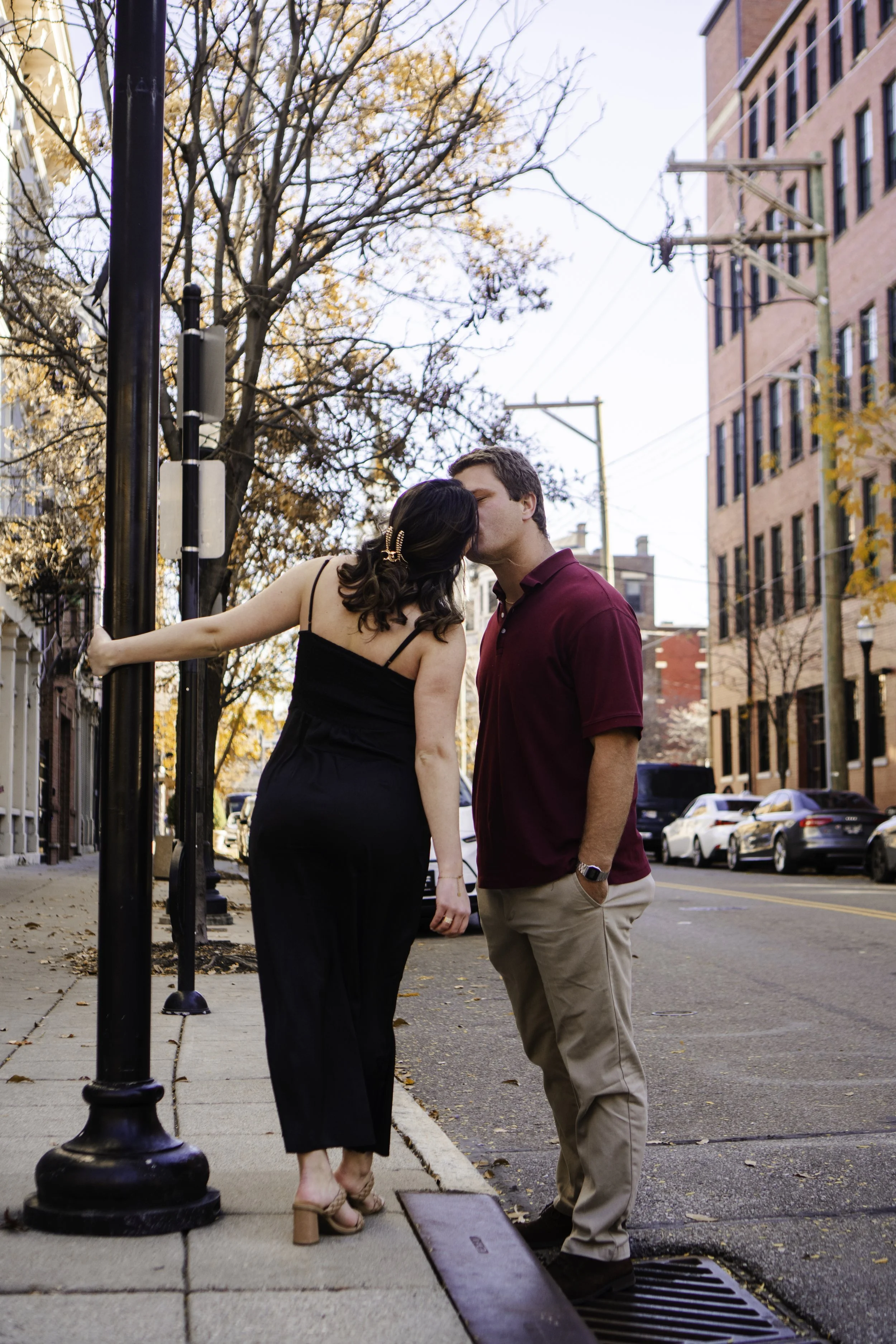 A couple sharing a kiss on a city sidewalk during fall, with autumn leaves on trees and parked cars along the street.