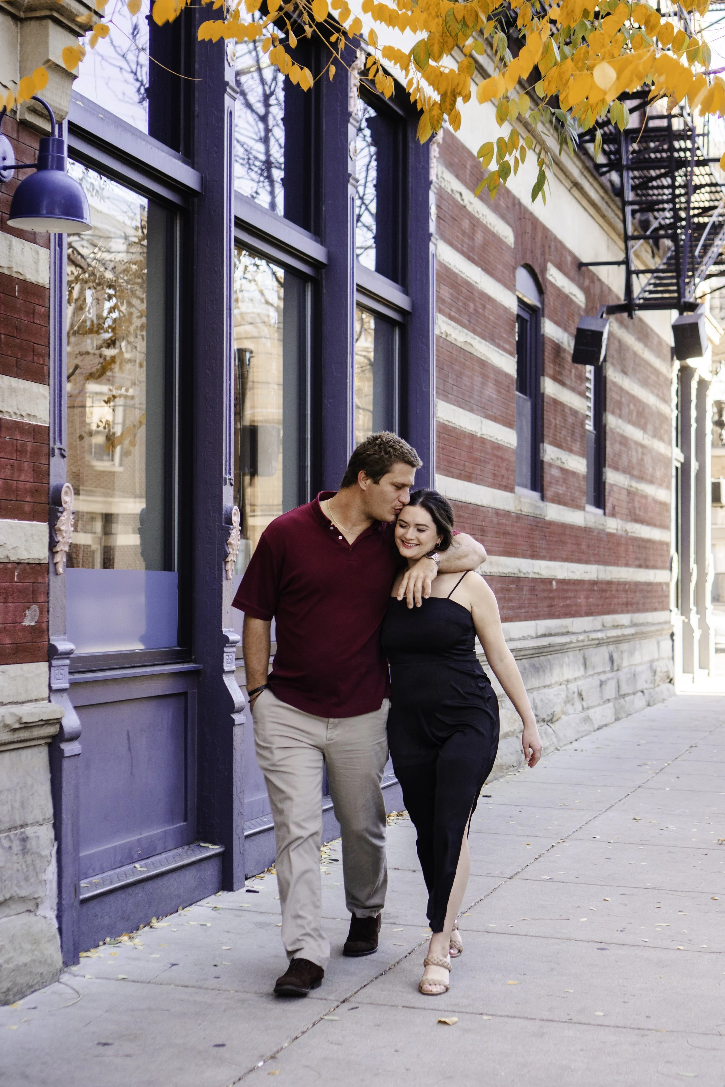 A young couple walking arm-in-arm outside a brick building with blue window frames, autumn leaves overhead, and sunlight casting shadows on the sidewalk.