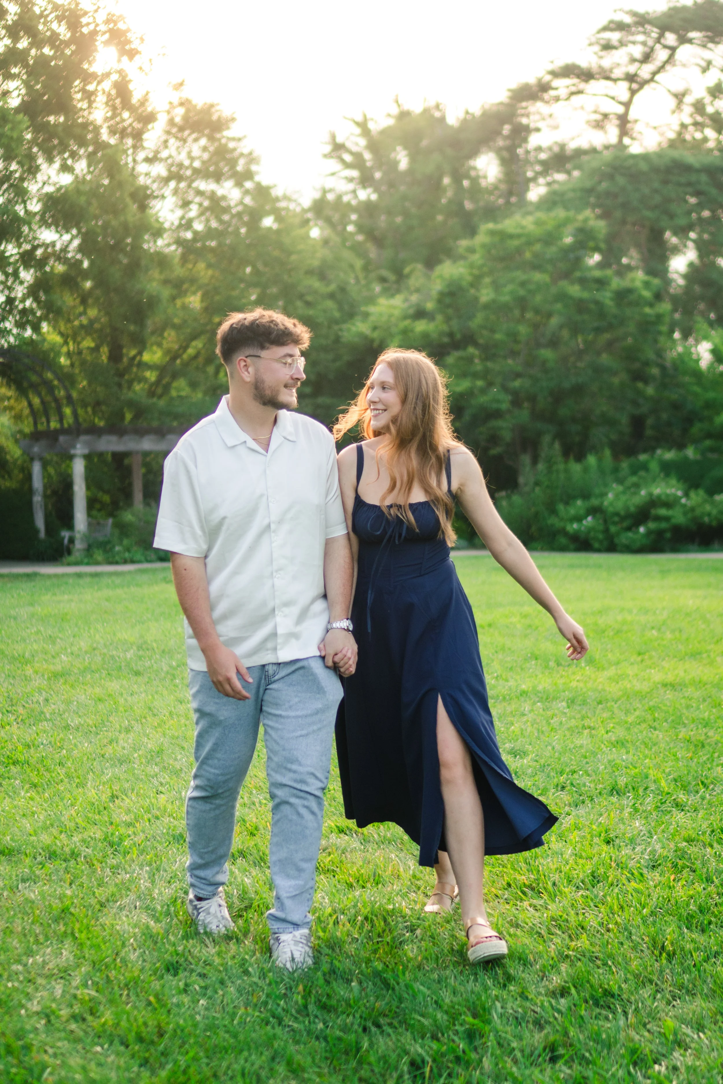 A young couple walking outdoors on a grassy field, holding hands, smiling, with trees and a pergola in the background, during sunset.