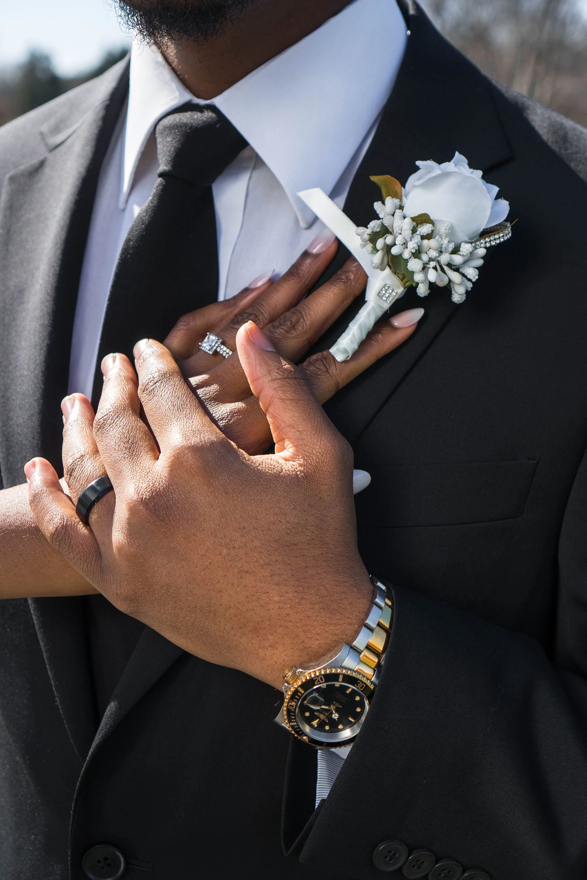 Close-up of a person in a black suit and white shirt, with their hand holding a white boutonniere with a white rose and small white flowers, pinned to their lapel. The person is wearing a gold and silver watch and a black ring on their finger, and the background shows an outdoor setting.