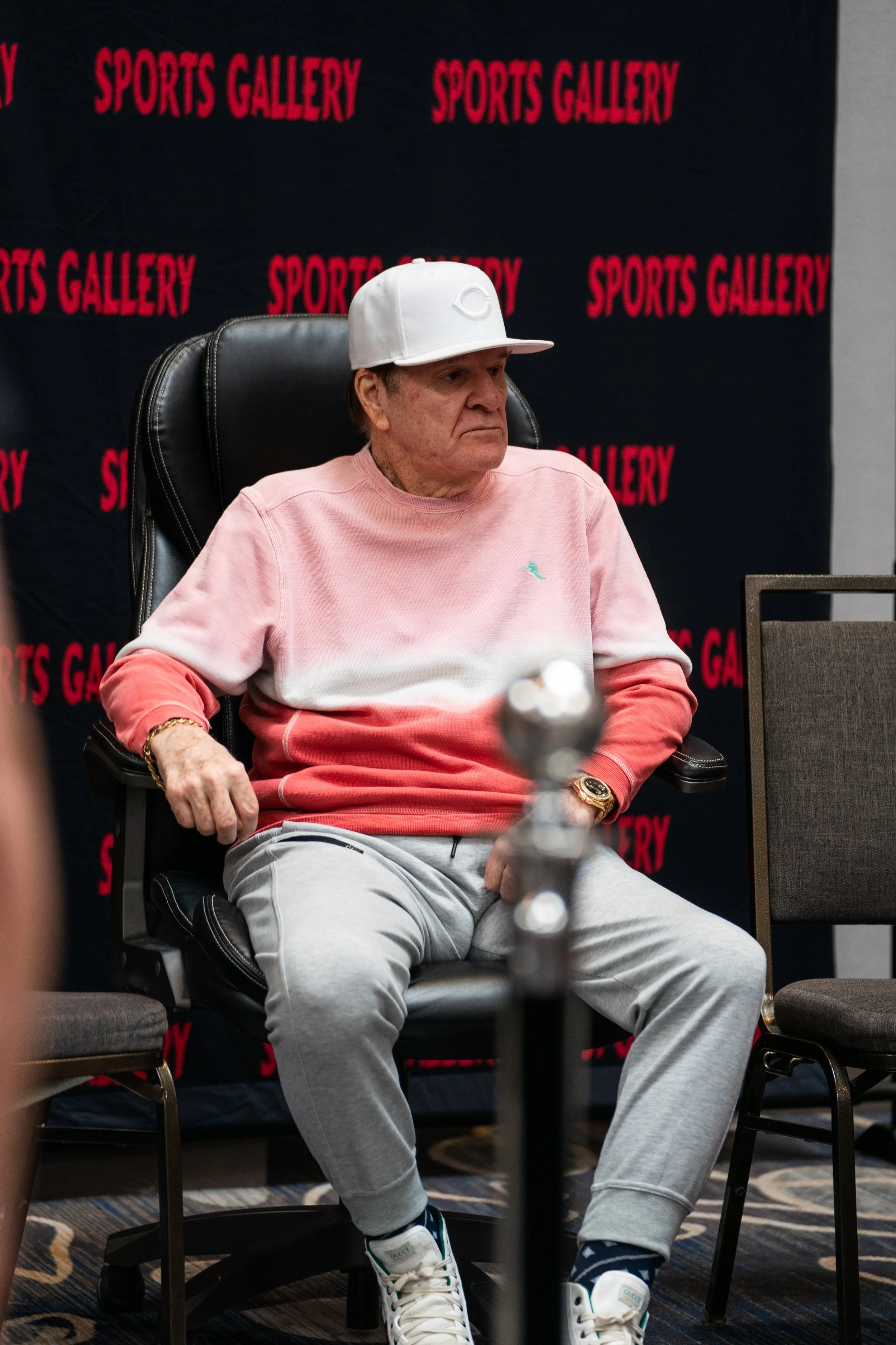 Older man sitting in a black leather office chair at a Sports Gallery event. He wears a white cap, pink gradient sweatshirt, gray sweatpants, and sneakers. There is a black backdrop with red "Sports Gallery" text behind him.