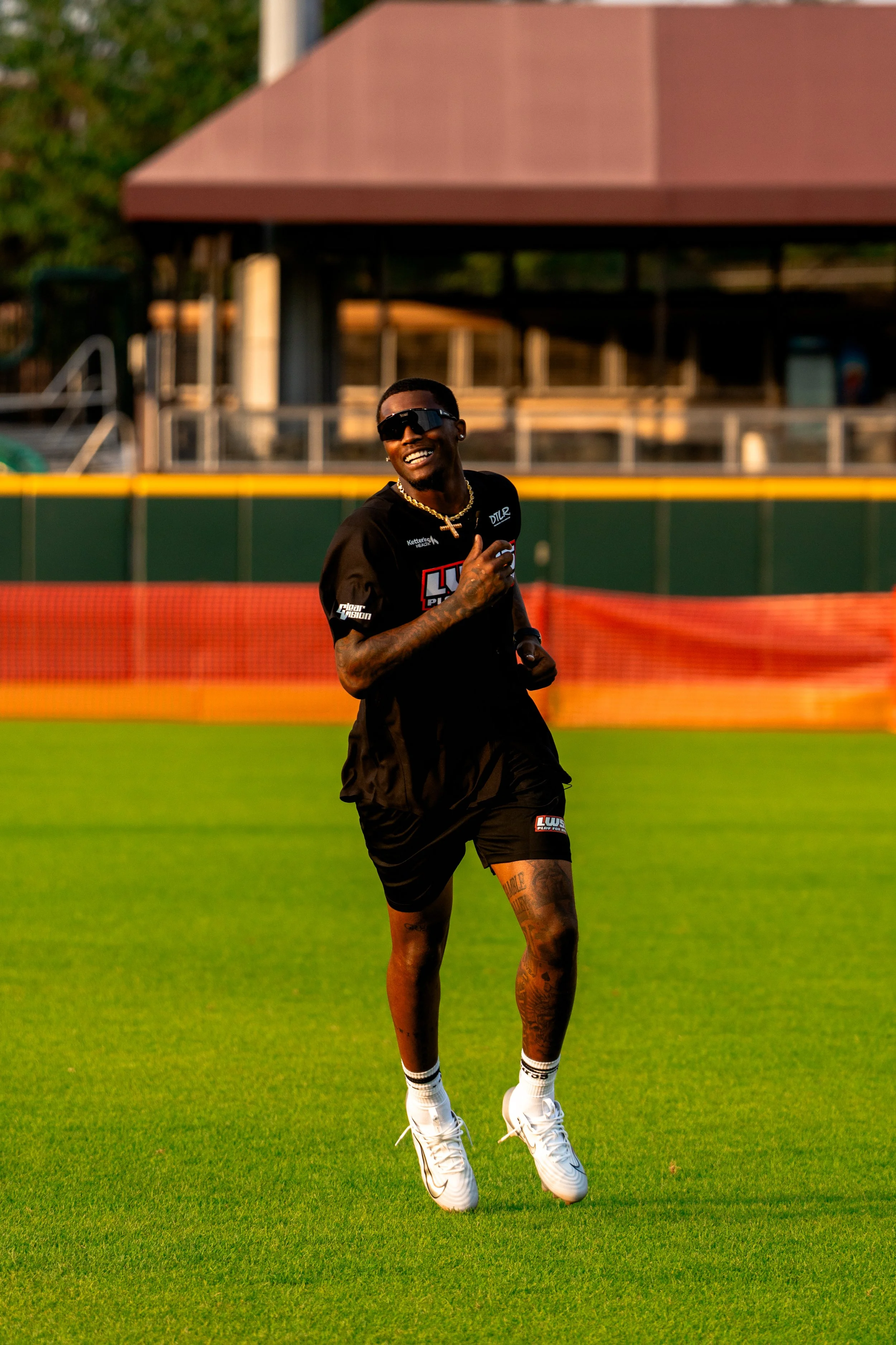 A man in black sports attire, with tattoos, white athletic shoes, sunglasses, and a gold chain, is smiling and running on a baseball or football field during daytime.