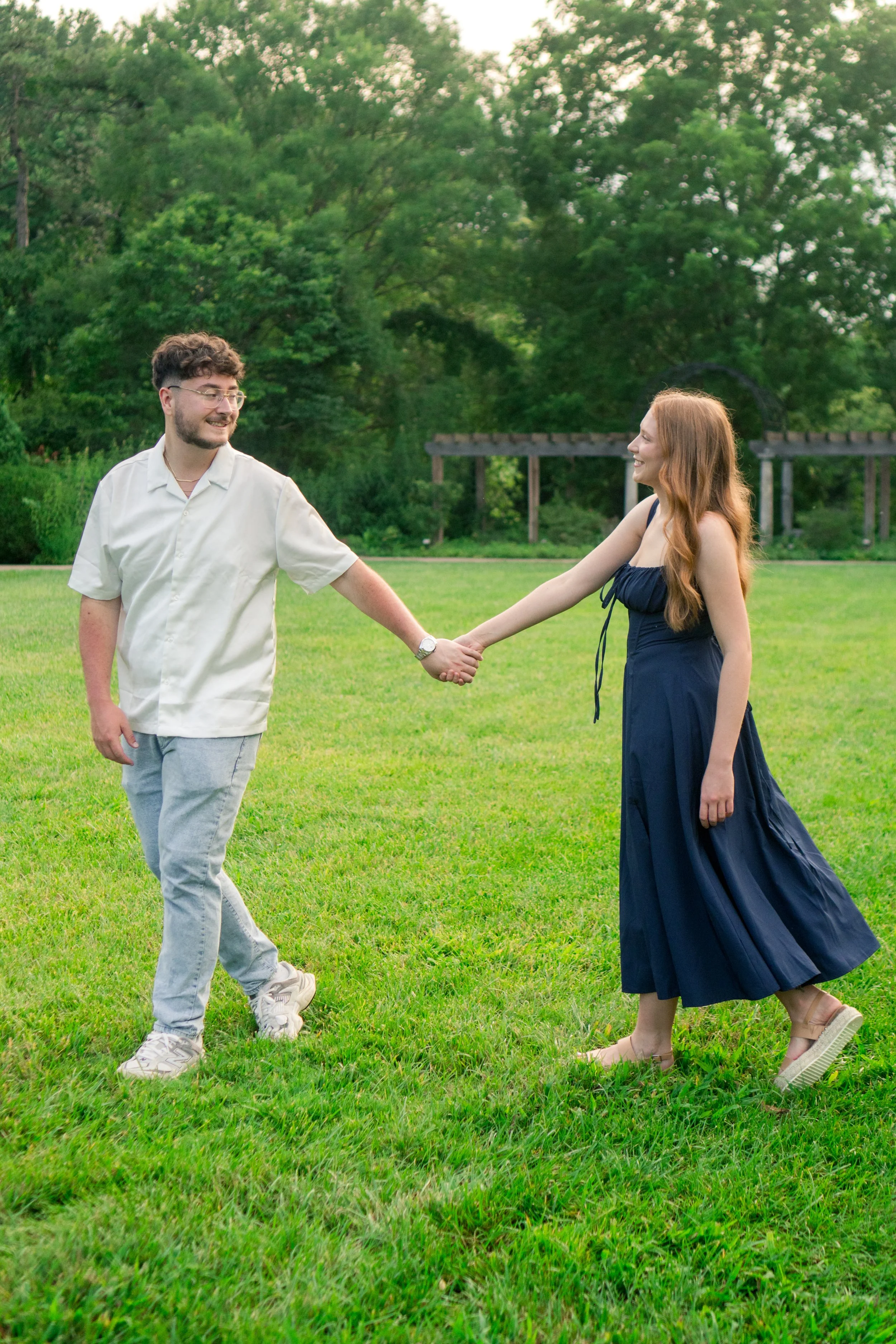 A man and a woman holding hands in a grassy park, smiling at each other. The man wears glasses, a short-sleeved white shirt, and light gray pants; the woman wears a navy blue dress and beige platform sandals. Green trees and a wooden structure are in the background.