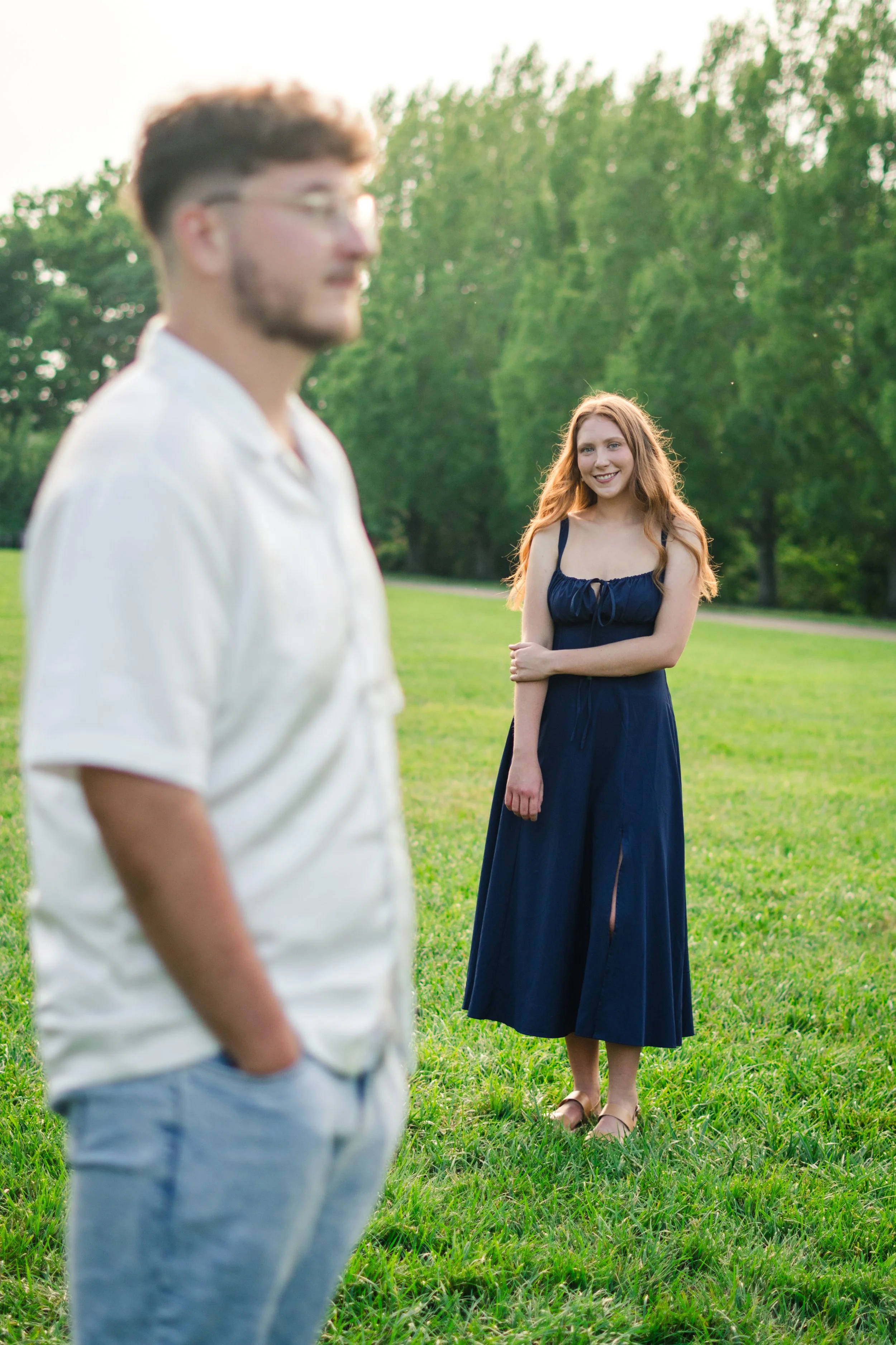 A young man in a white shirt and light blue jeans stands in the foreground with hands in pockets, slightly out of focus. In the background, a smiling young woman with long red hair, wearing a dark blue dress and sandals, stands on a grassy field with
