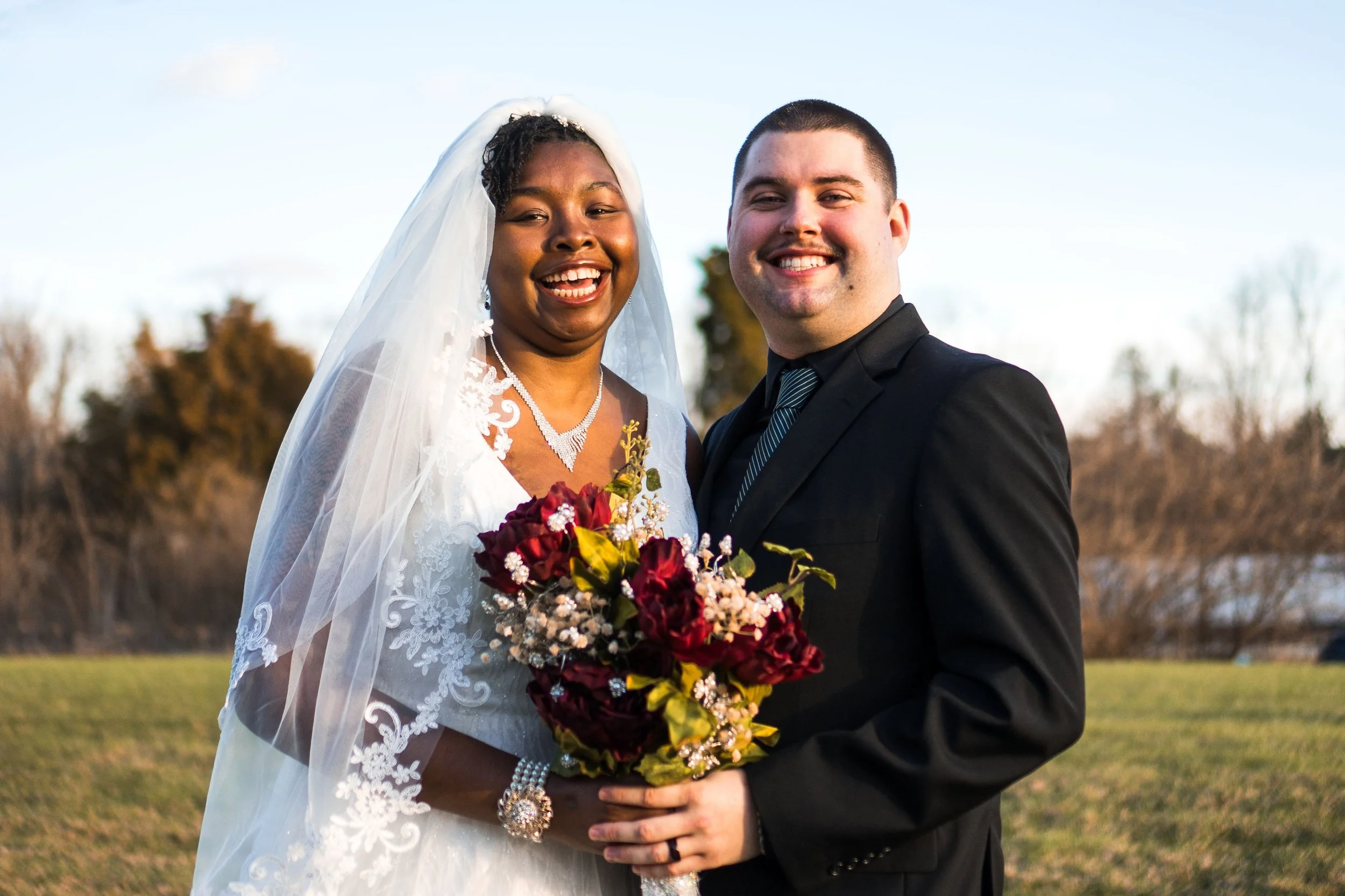 A smiling bride and groom standing outdoors with a bouquet of flowers, dressed in wedding attire, during daytime.