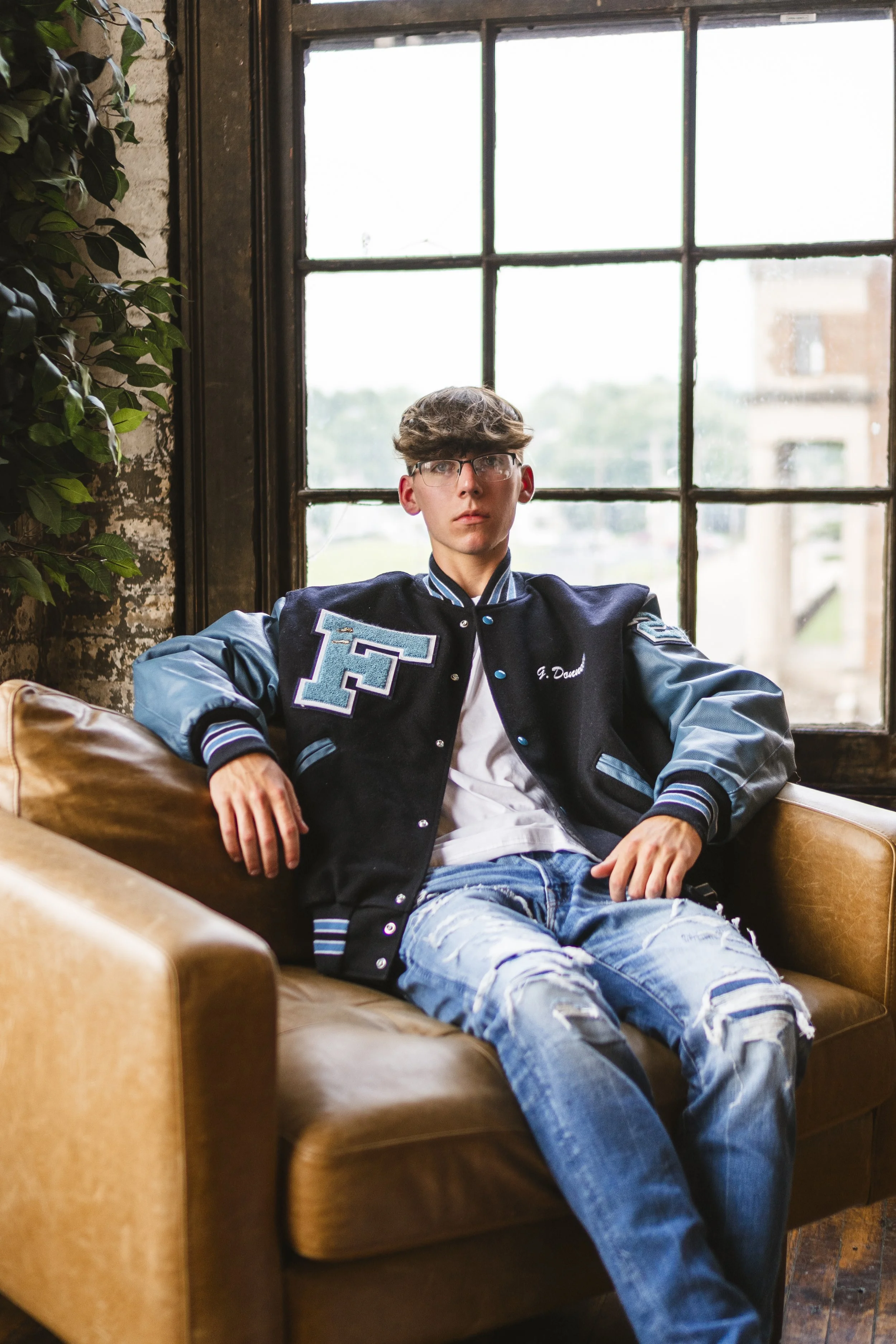 A young man with glasses and light brown hair sitting on a tan leather sofa in front of a large window. He is wearing a black and blue varsity jacket with a letter 'F' on the front and the name 'G. Douse' on it, along with ripped blue jeans and a whi