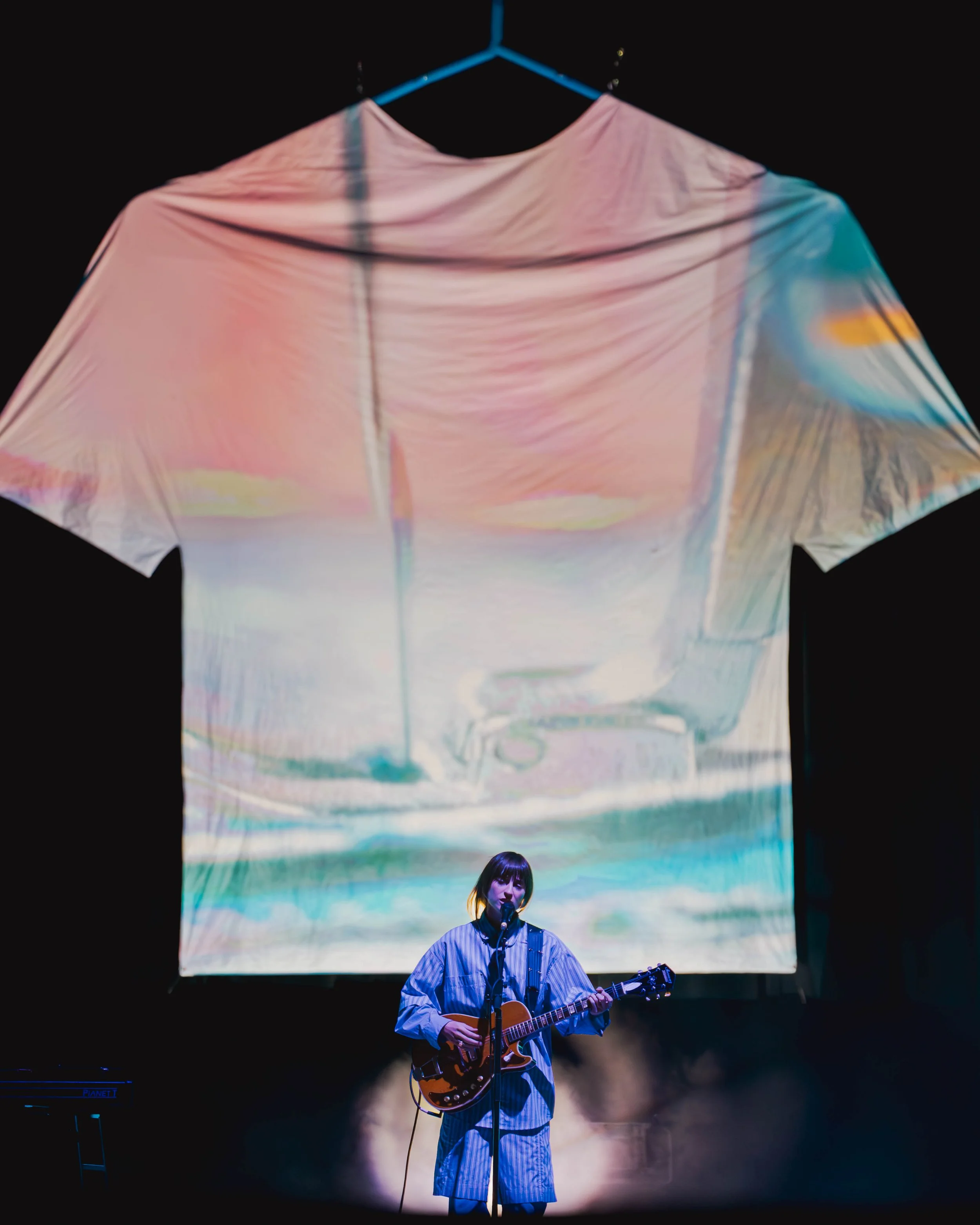A woman performing on stage with a guitar, standing in front of a large screen with a colorful, abstract projection.