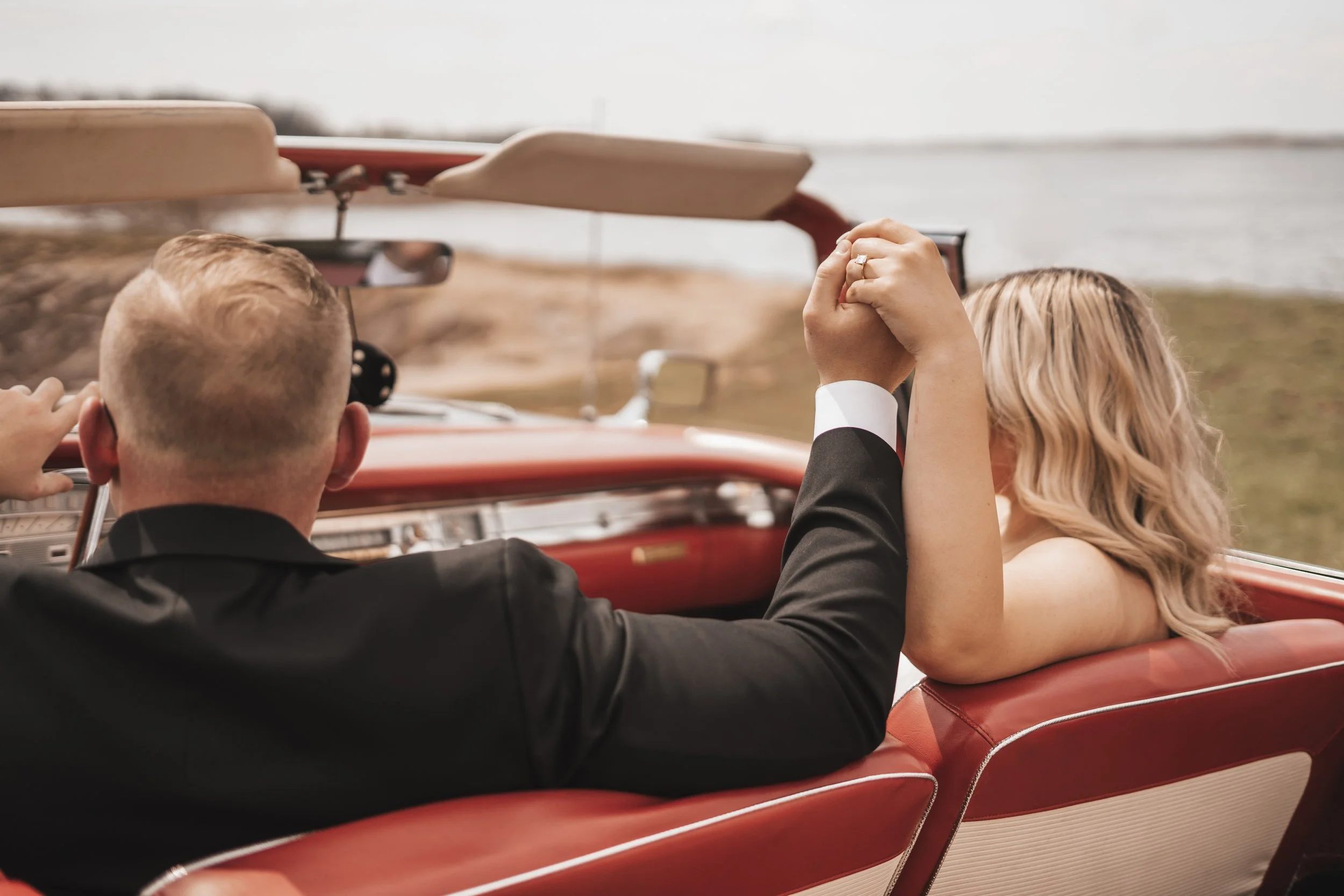 A couple holding hands in a vintage red convertible car, with a water body and landscape in the background.