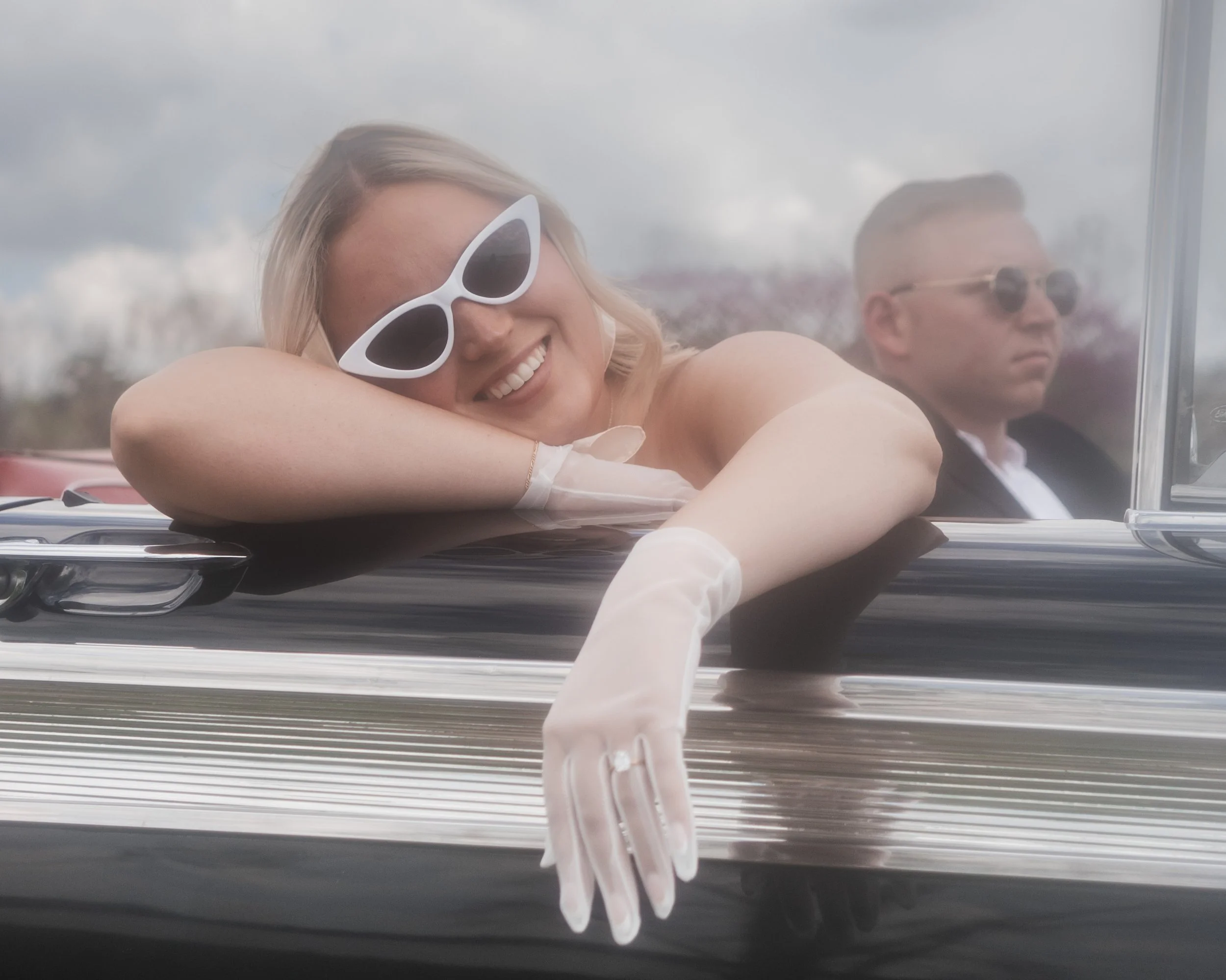 A woman with blonde hair smiling and wearing white sunglasses relaxing in a black convertible car with her arm resting on the door, and a man with short hair and sunglasses sitting in the background.