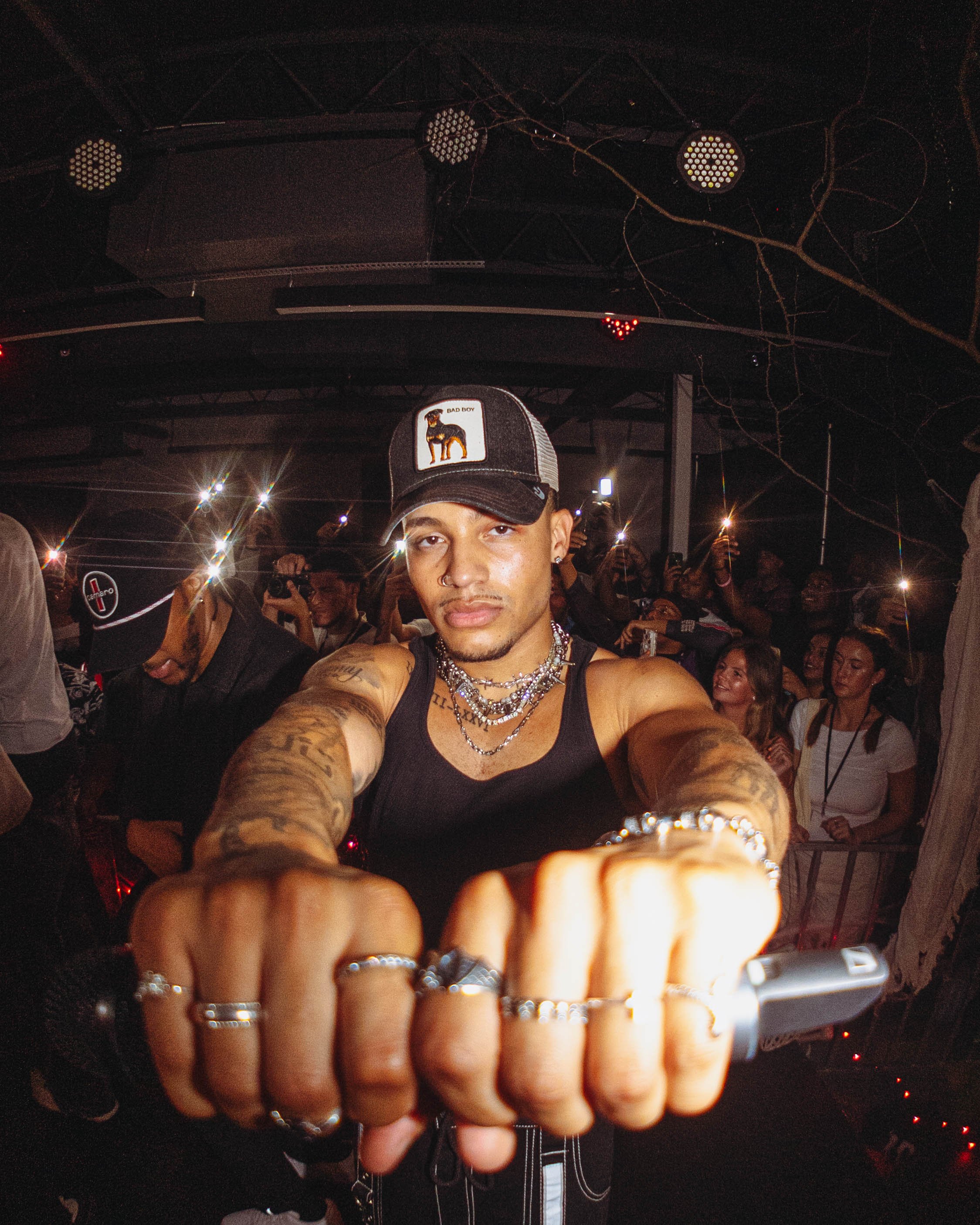 A young man with tattoos taking a selfie at a crowded nightclub, wearing a black tank top, multiple jewelry pieces, and a trucker cap with a dog patch.