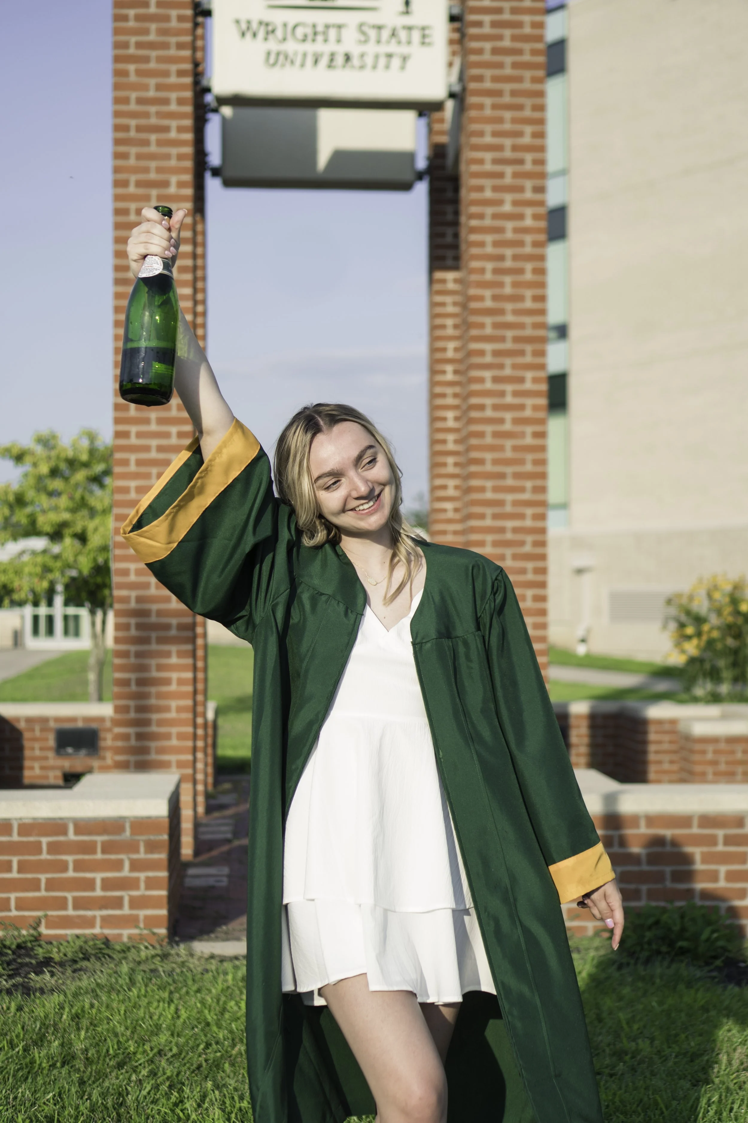 A young woman wearing a green graduation gown and cap, holding a green bottle, celebrating outside a brick building with a sign that reads "Wright State University."