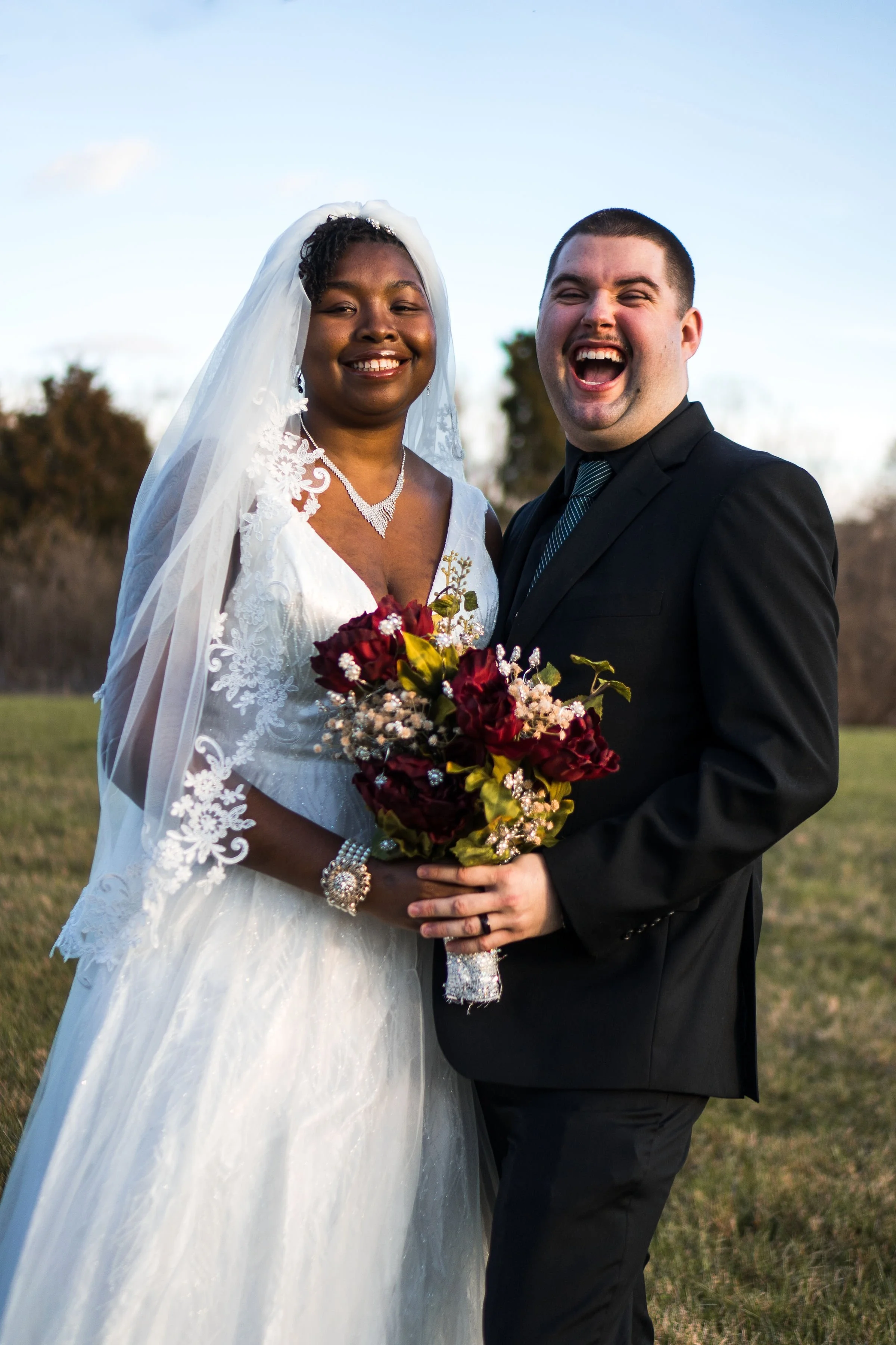A joyful bride and groom standing outdoors on a grassy field, smiling and holding a bouquet of dark red and pink flowers. The bride wears a white lace wedding dress with a veil, and the groom wears a black suit with a striped tie.