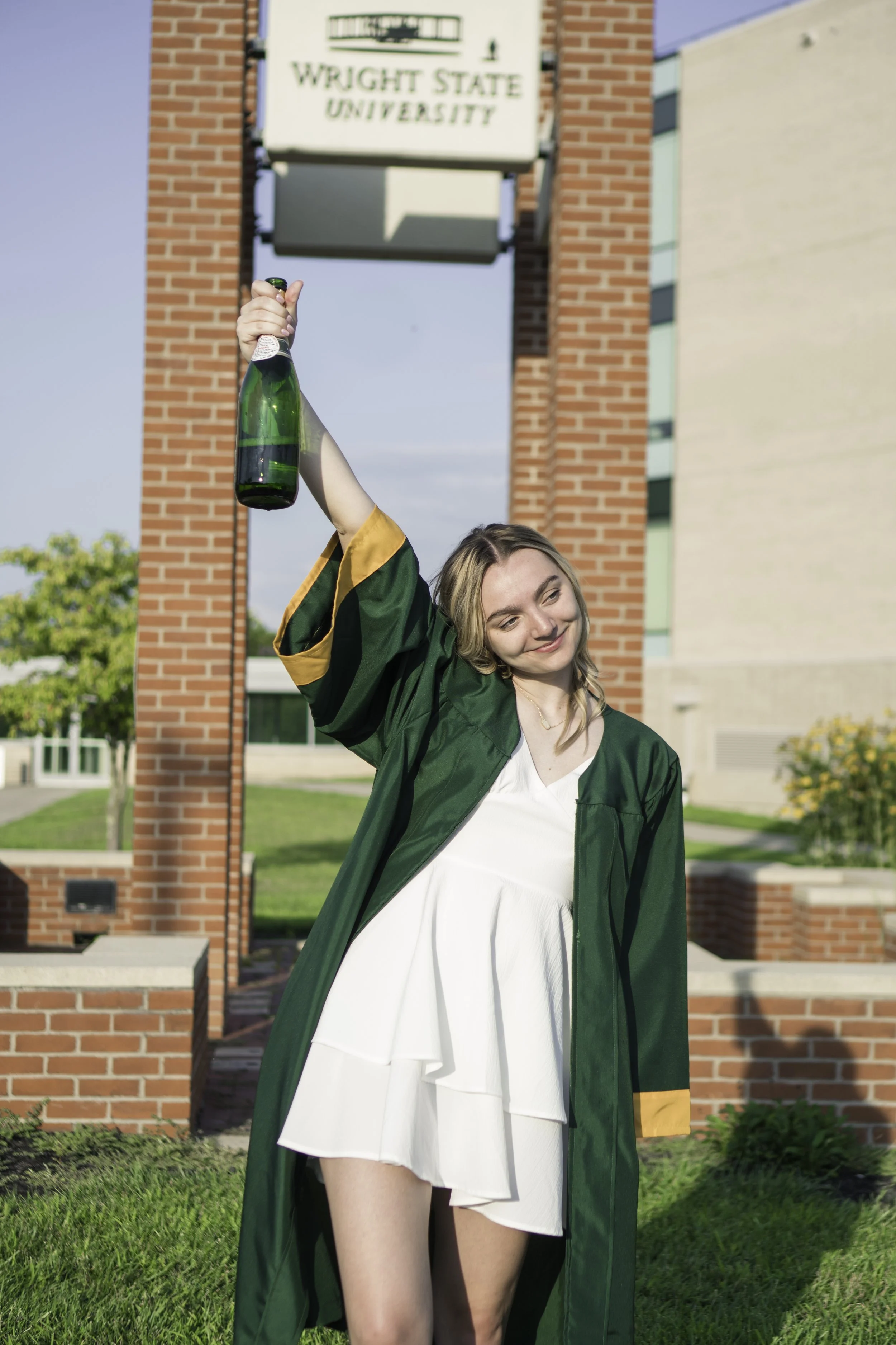 A young woman in a graduation gown holding a champagne bottle in the air outside of Wright State University.