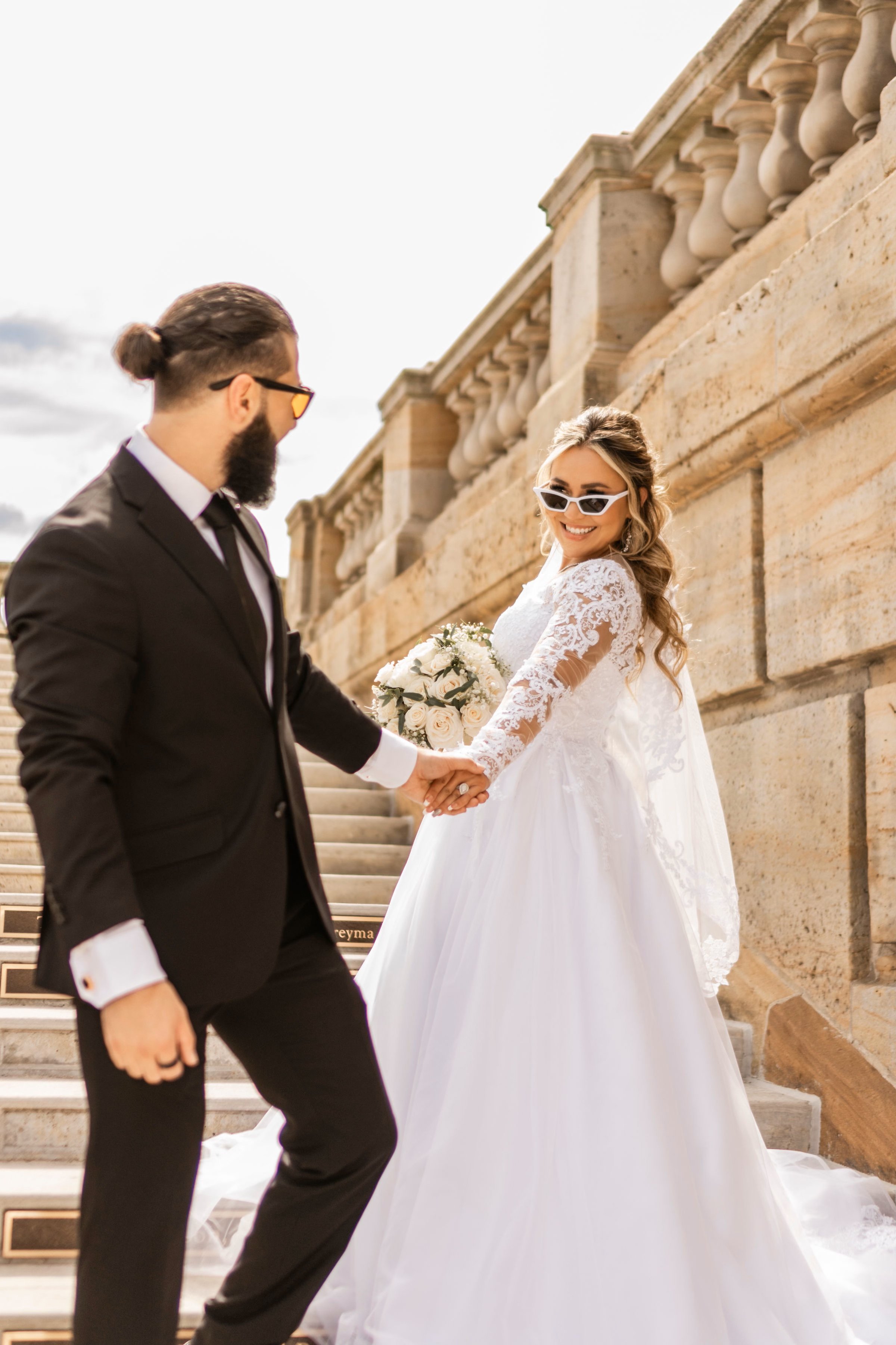 A bride and groom holding hands, smiling, outdoors on a staircase with stone railings, the bride in a white lace wedding gown with long sleeves and sunglasses, the groom in a black tuxedo with a black tie, she holding a bouquet of roses.