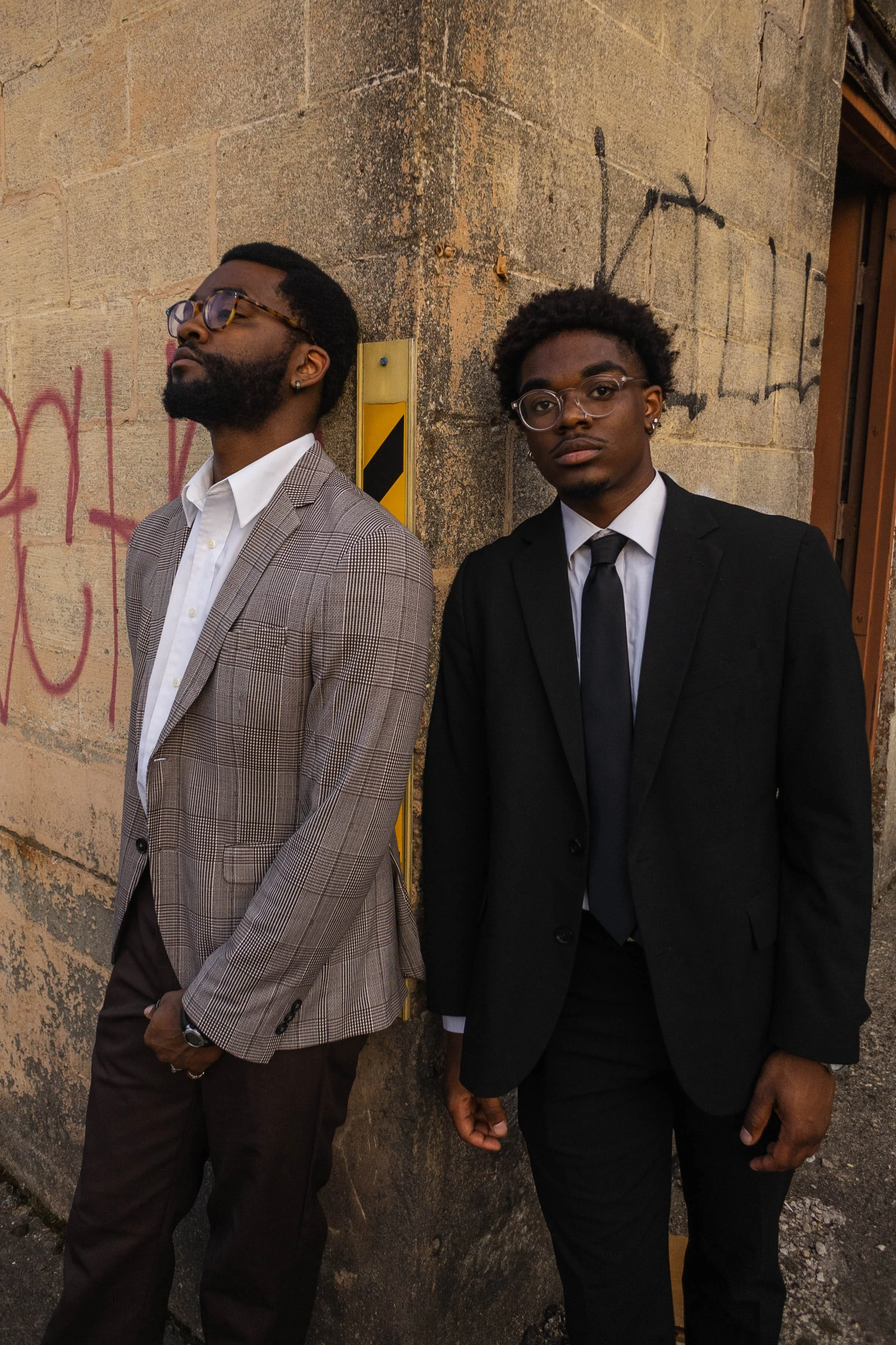 Two men in formal attire standing against a graffiti-covered brick wall. The man on the left is wearing a plaid blazer, white shirt, glasses, and has facial hair, with his eyes closed. The man on the right is wearing a black suit, white shirt, black 